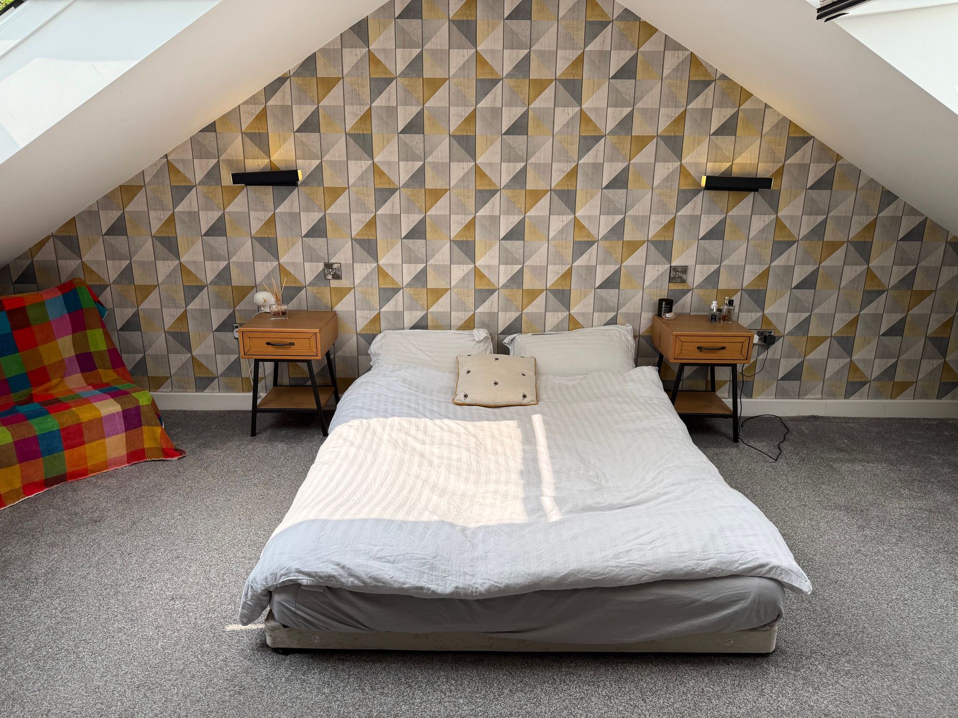 Bedroom with geometric wallpaper, low bed, two bedside tables, and a colorful blanket.