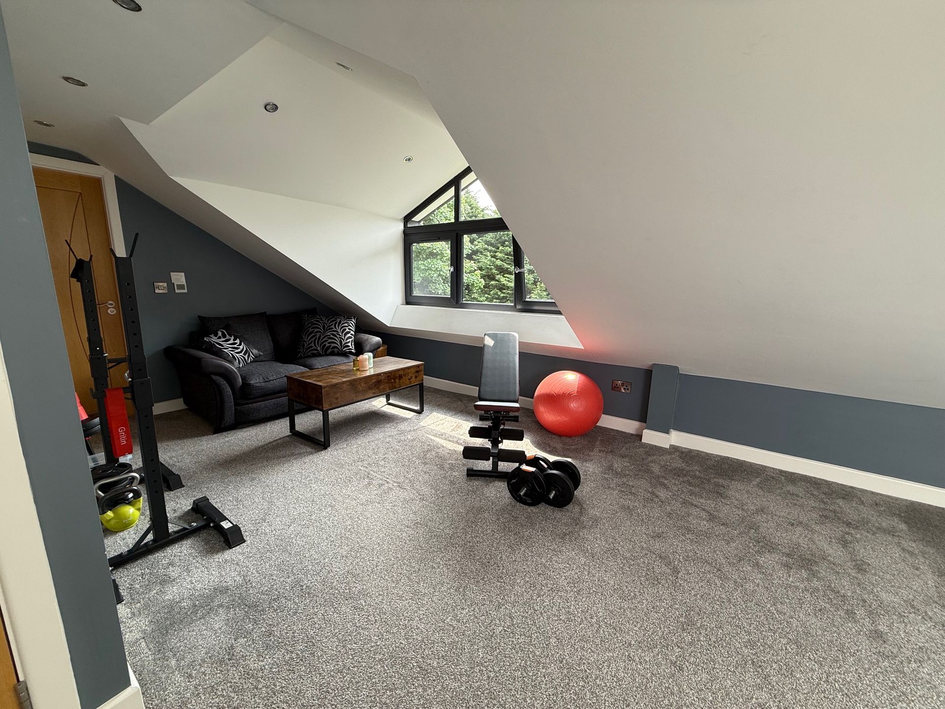 Attic home gym with gray carpet, blue walls, a black couch, weights, and a red exercise ball.