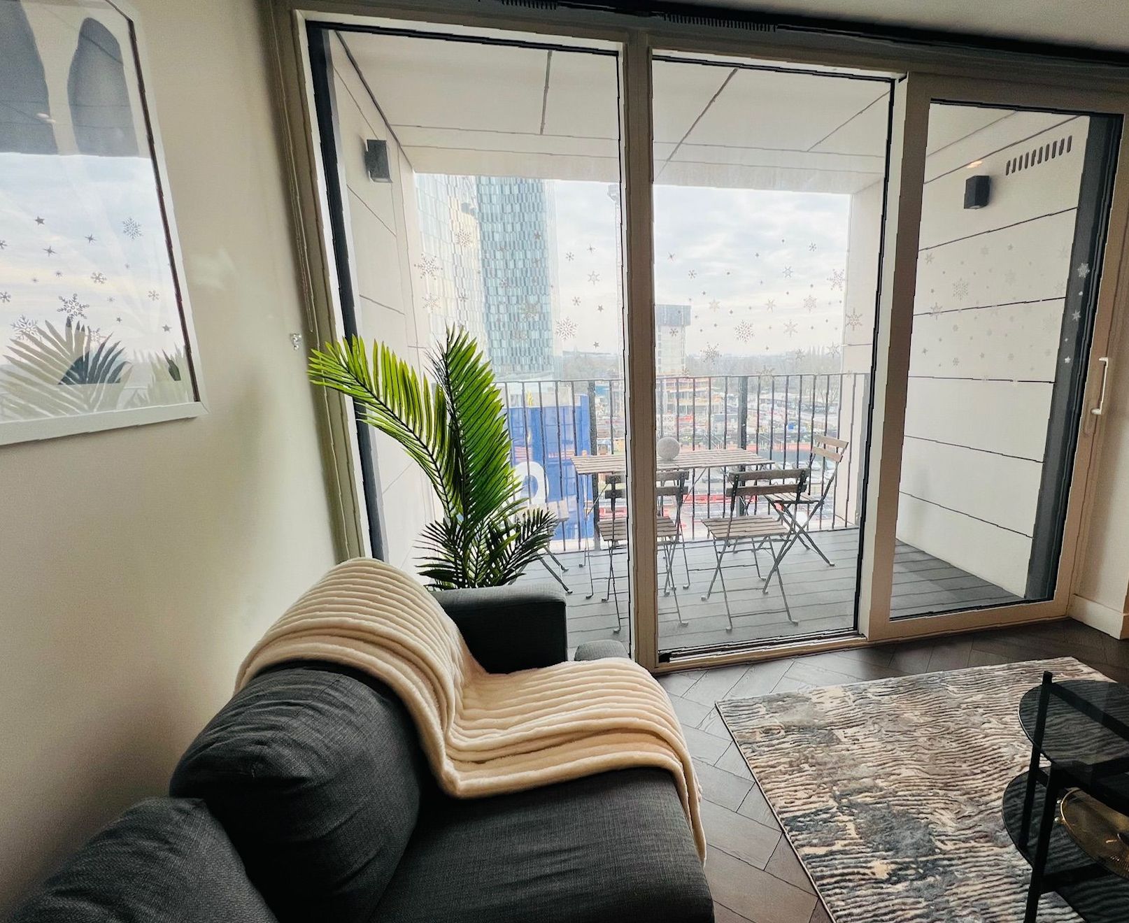 Cozy living room with grey couch, blanket, and balcony with city view. Plant and rug included.