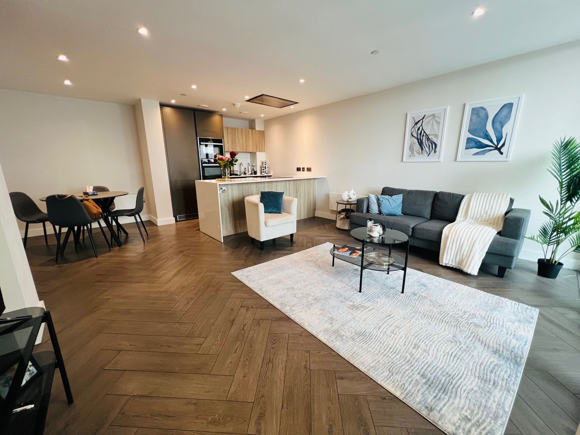 Open-concept living space with dark wood floors, grey sofa, kitchen, dining table, and rug.