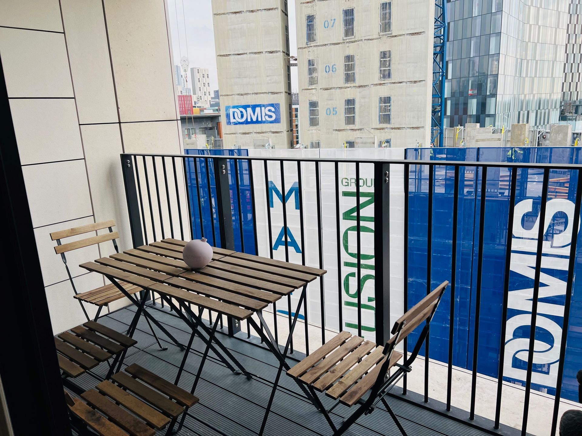 Small balcony with a wooden table, two chairs, and a decorative orb. Blue construction site visible in the background.