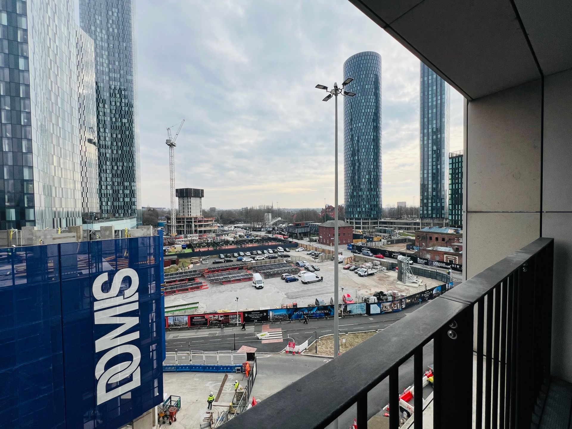 Construction site viewed from balcony, tall buildings in background. Blue and white Domis hoarding visible.