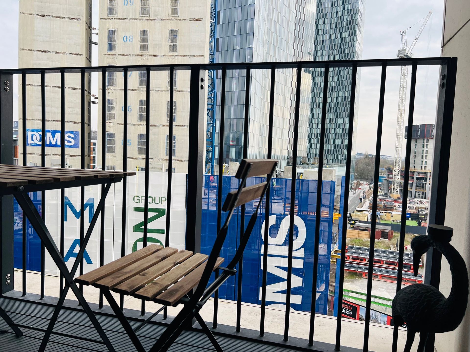 Balcony with small table, chair, and decorative bird overlooking cityscape with construction.