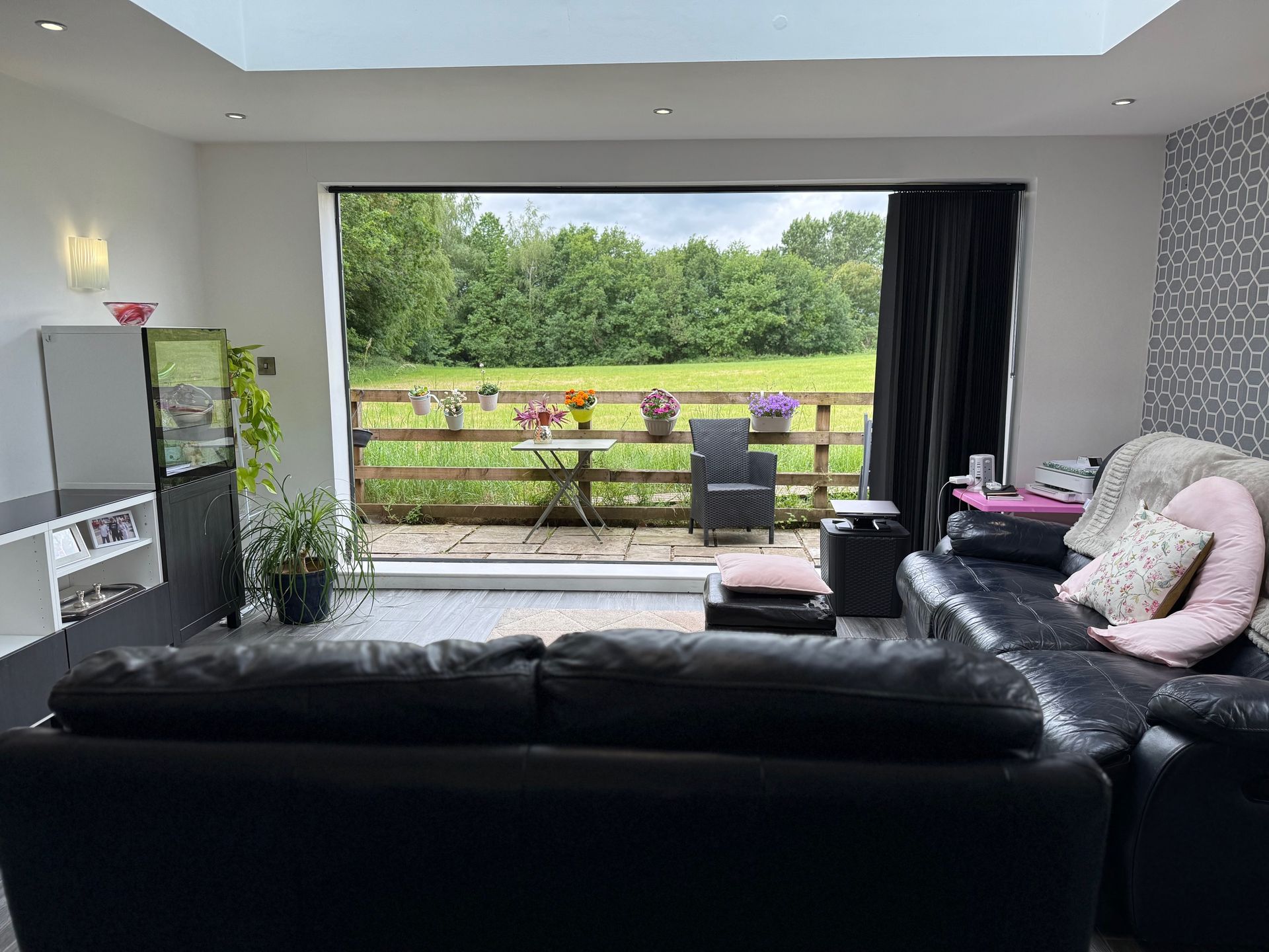 Living room with a black sectional sofa, open to a patio with a table and chairs, overlooking a grassy area.