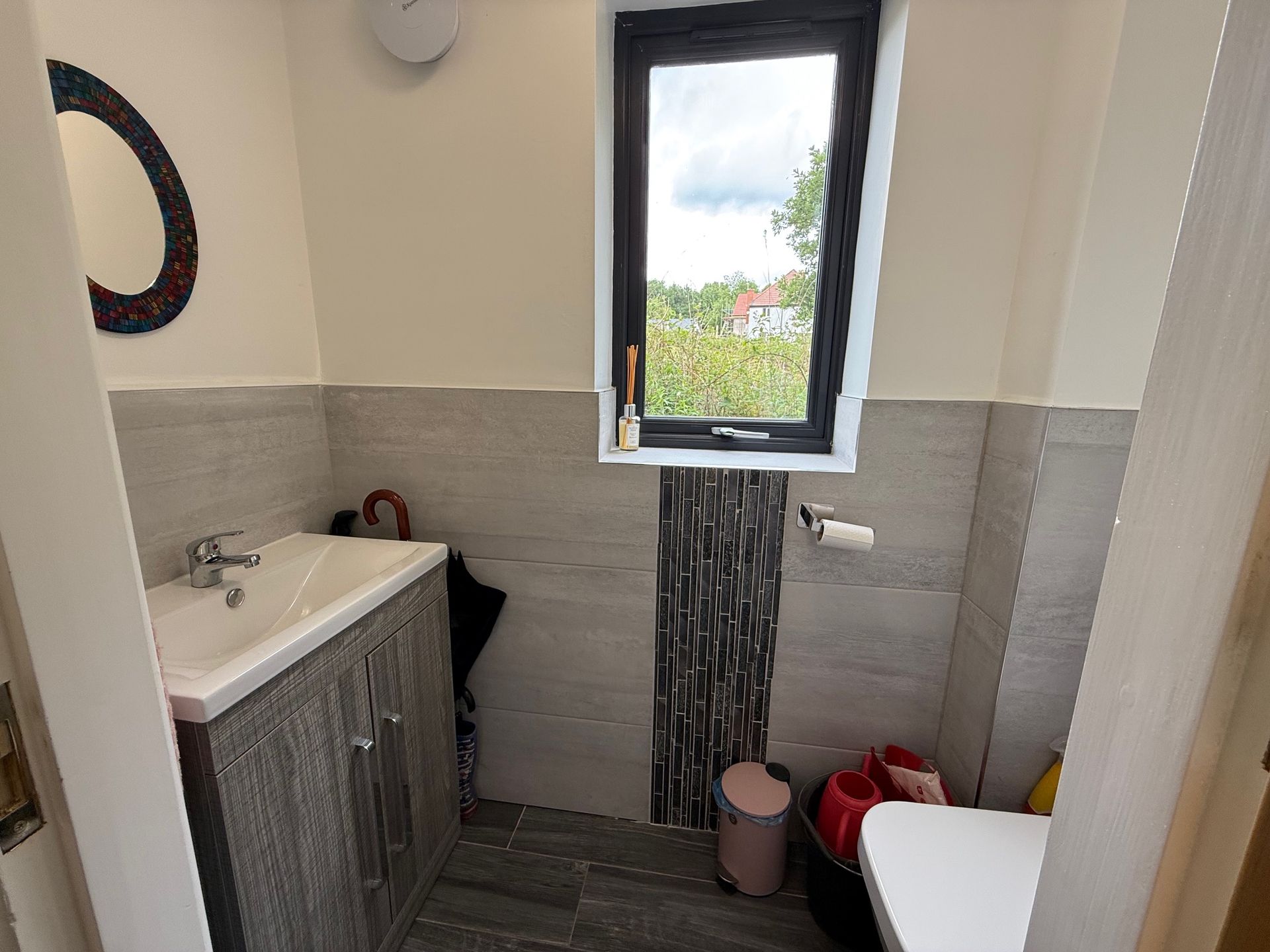 Small bathroom with a sink, toilet, and window, neutral tones, gray tile, and a decorative mosaic mirror.