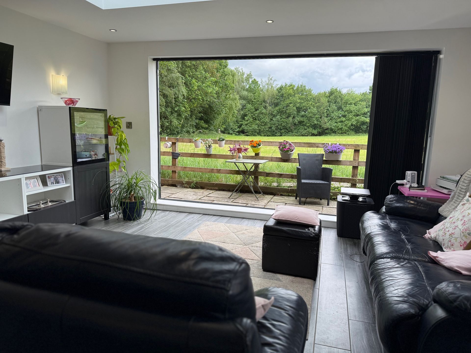 Living room with large window overlooking a garden and patio; black leather sofa, dark floors, neutral walls.