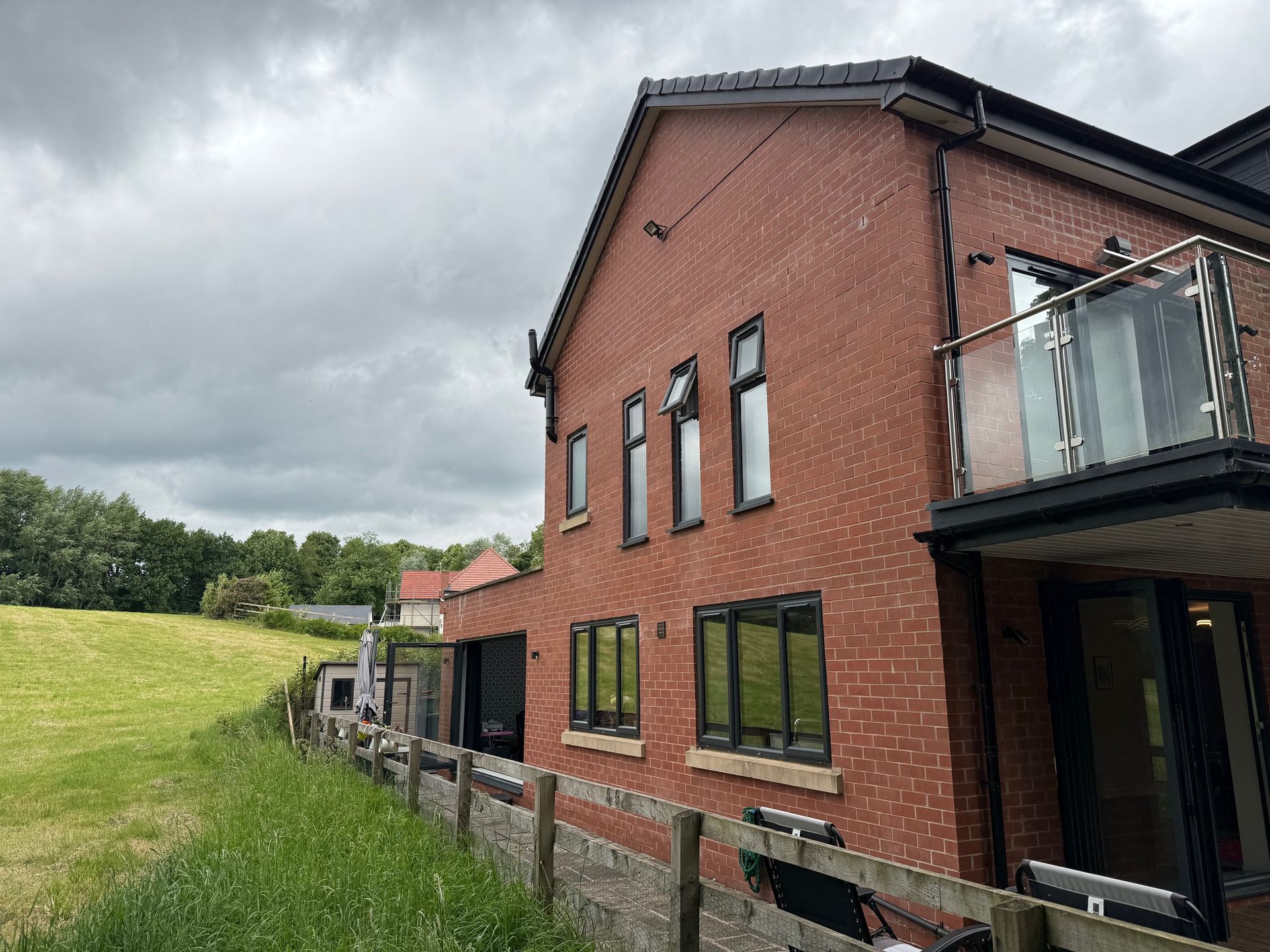 Red brick house with black-framed windows and a glass balcony, beside a grassy field under a cloudy sky.