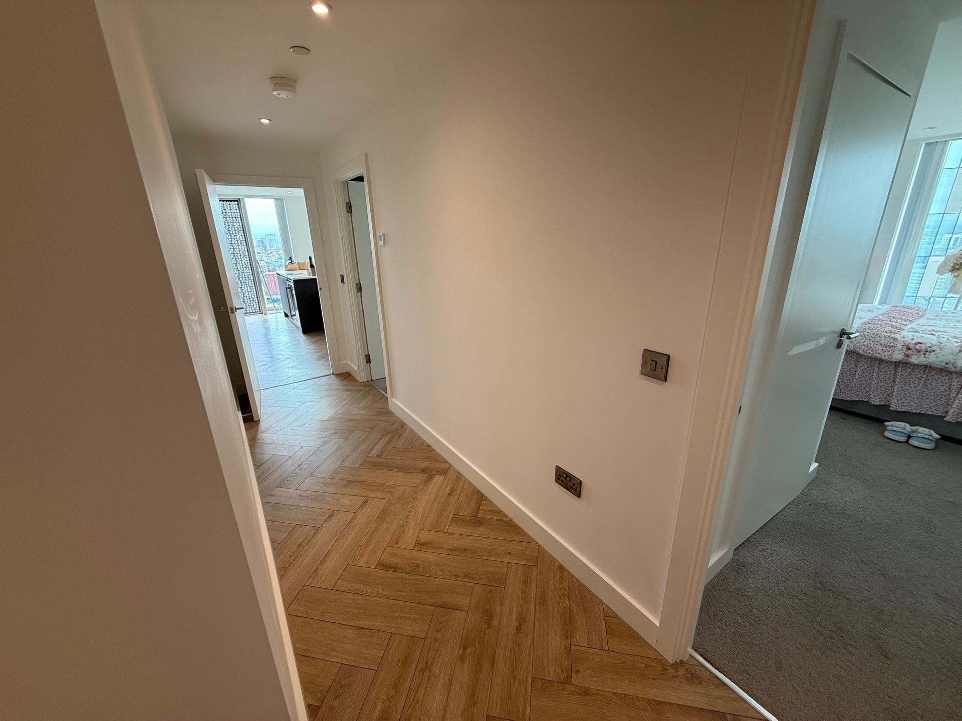 Hallway with herringbone wood flooring, white walls, and a bedroom visible on the right.