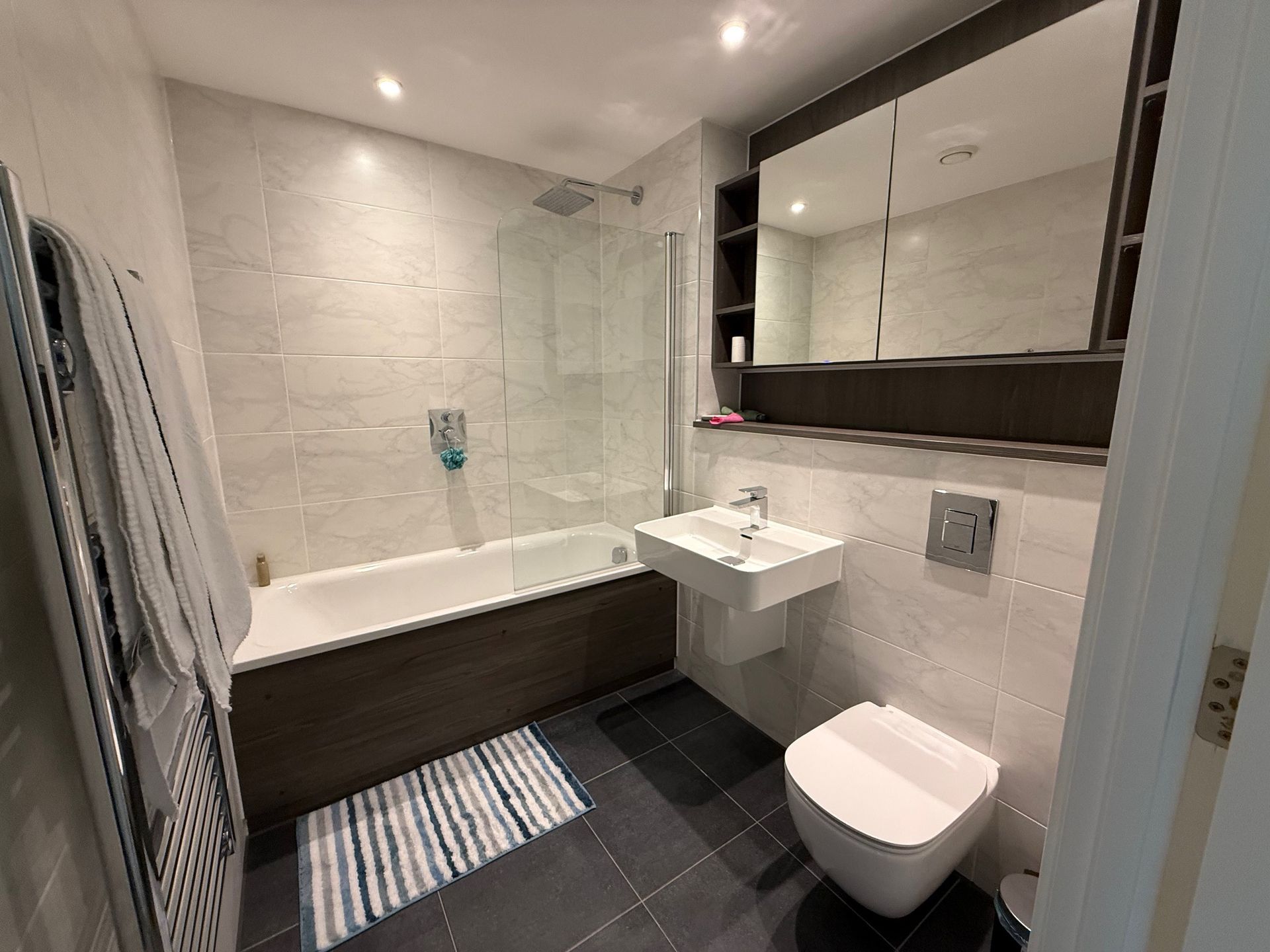 Bathroom with bathtub, sink, toilet, and large mirror cabinet; gray and white tile, dark floor.