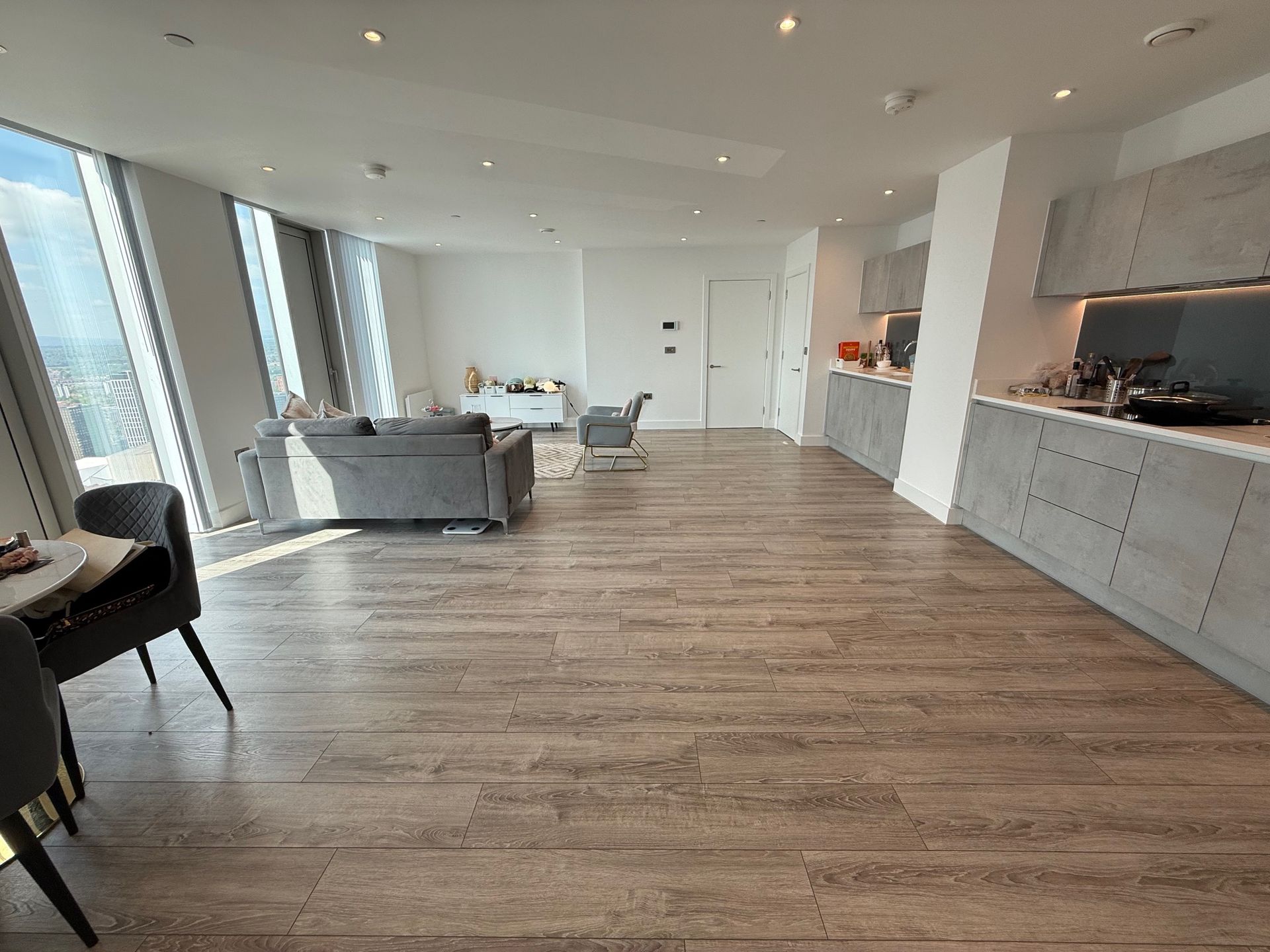 Spacious modern apartment interior with wooden floors, gray kitchen, and large windows.