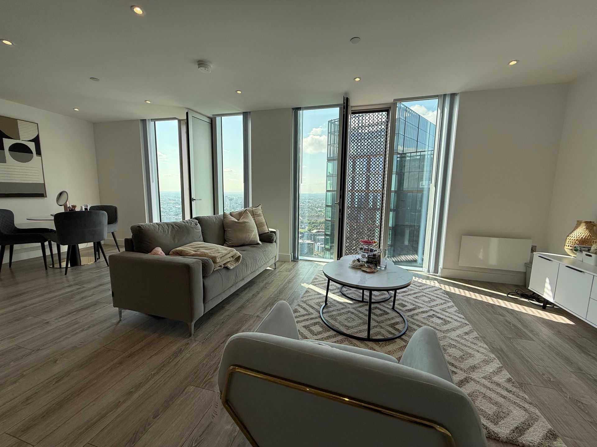 Living room with city view, grey sofa, armchair, coffee table, rug, dining table, and large windows.
