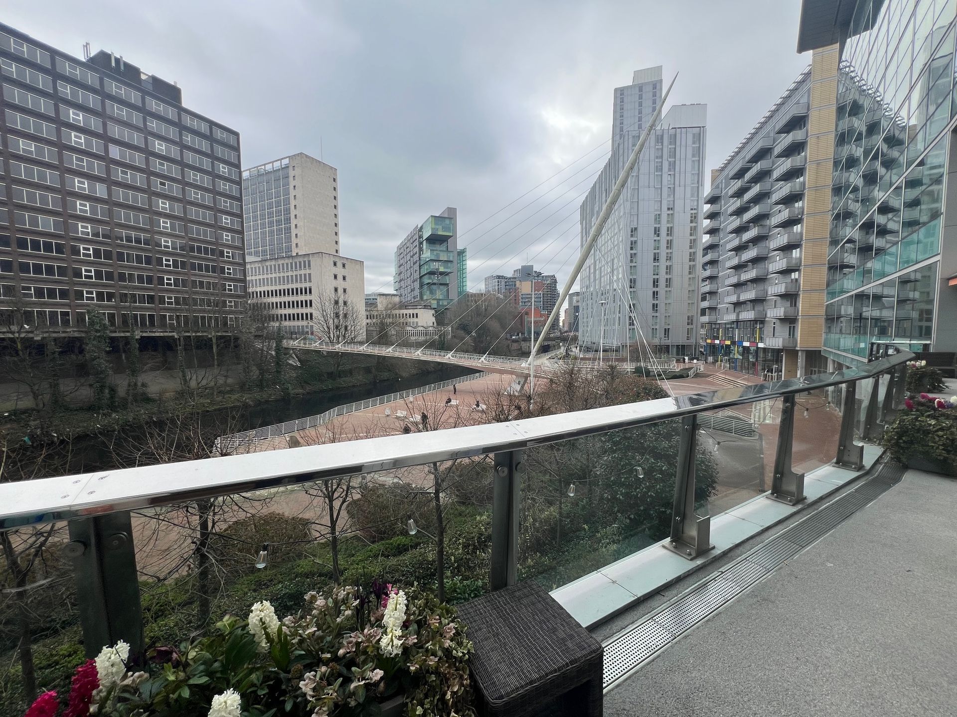 Cityscape with buildings overlooking a bridge, overcast sky. Railing and flowers in the foreground.