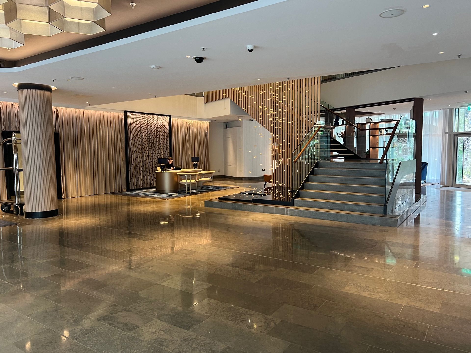 Hotel lobby: reception desk, staircase, polished floor, wooden accents, and an open doorway.