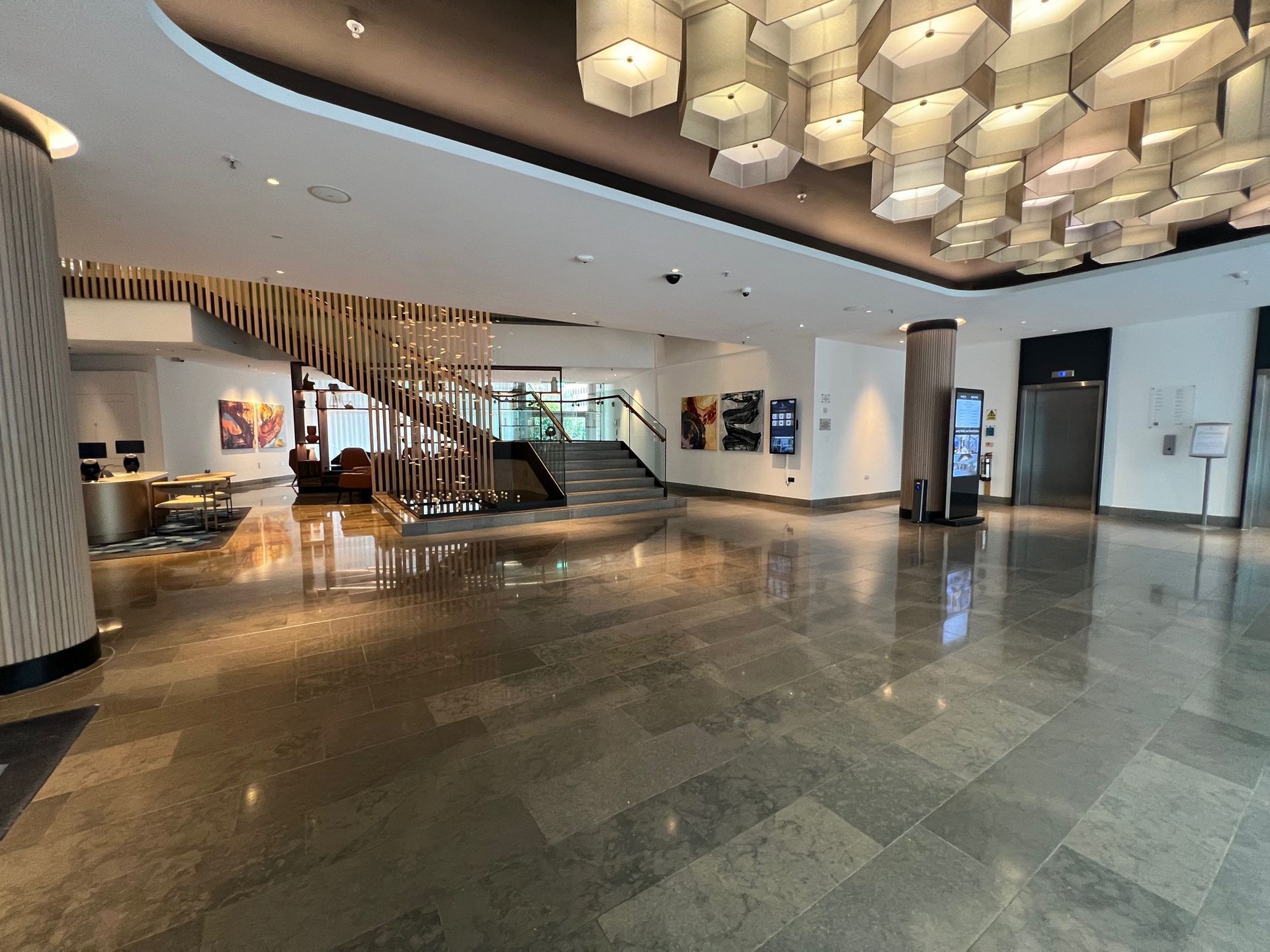 Hotel lobby with shiny tile floor, staircase, modern chandelier, and a digital information display.