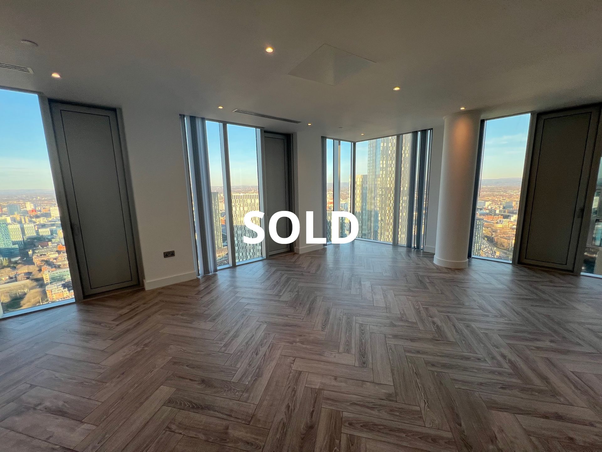 Spacious room with floor-to-ceiling windows and city views, herringbone floors. 