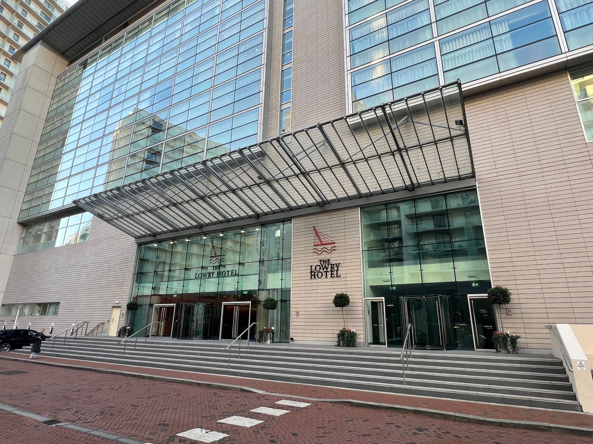 Exterior view of the Langham Hotel. Glass facade, entrance canopy, steps, red brick road, and cloudy sky.