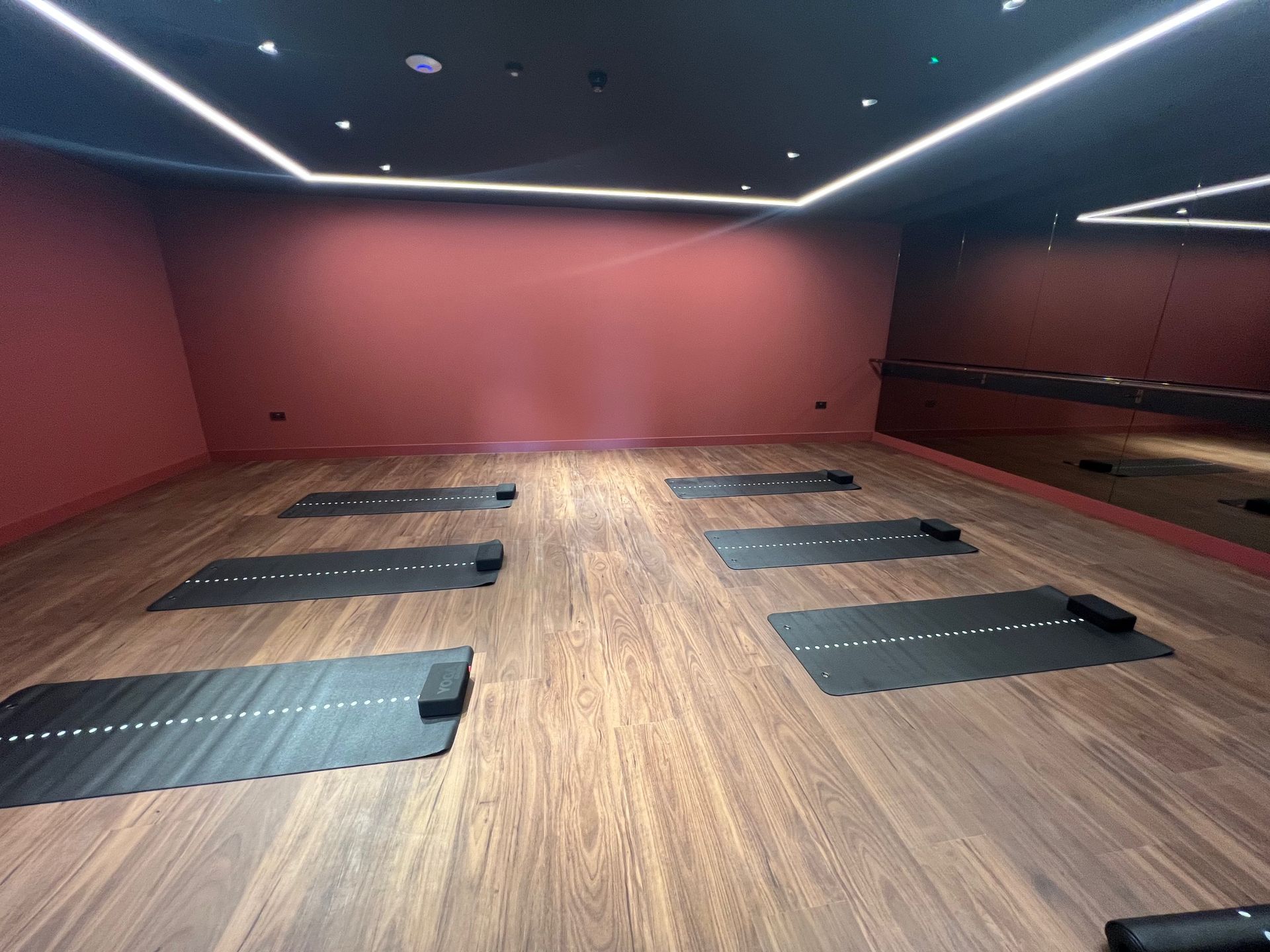 Yoga studio with black mats on wooden floor, red walls, mirror, and ceiling lights.