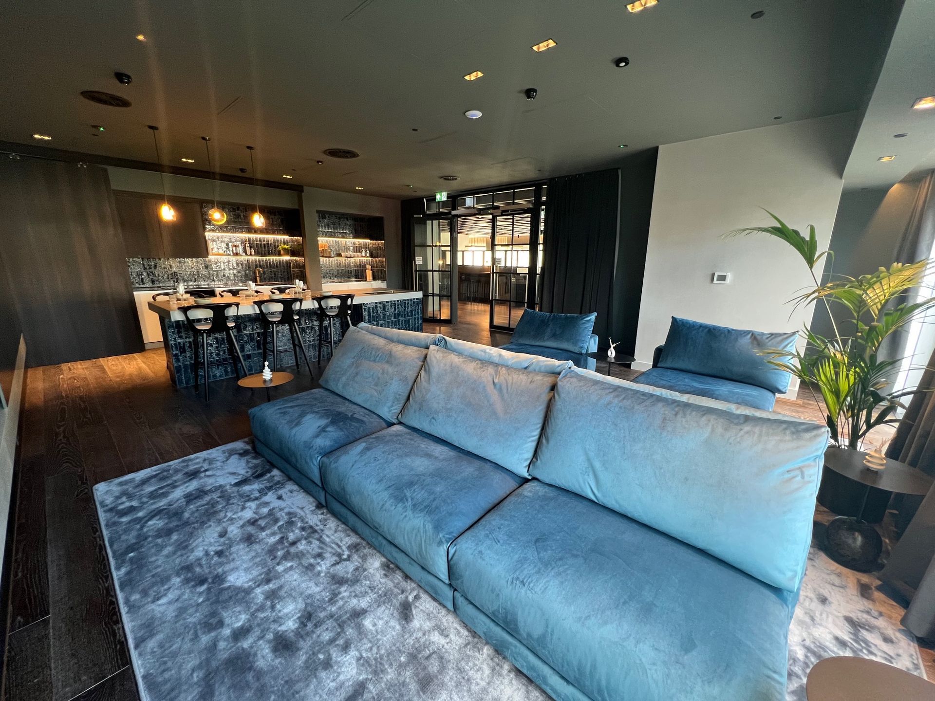 Lounge with a blue sofa, bar, and seating area. The setting has a modern design with dark walls and floors.