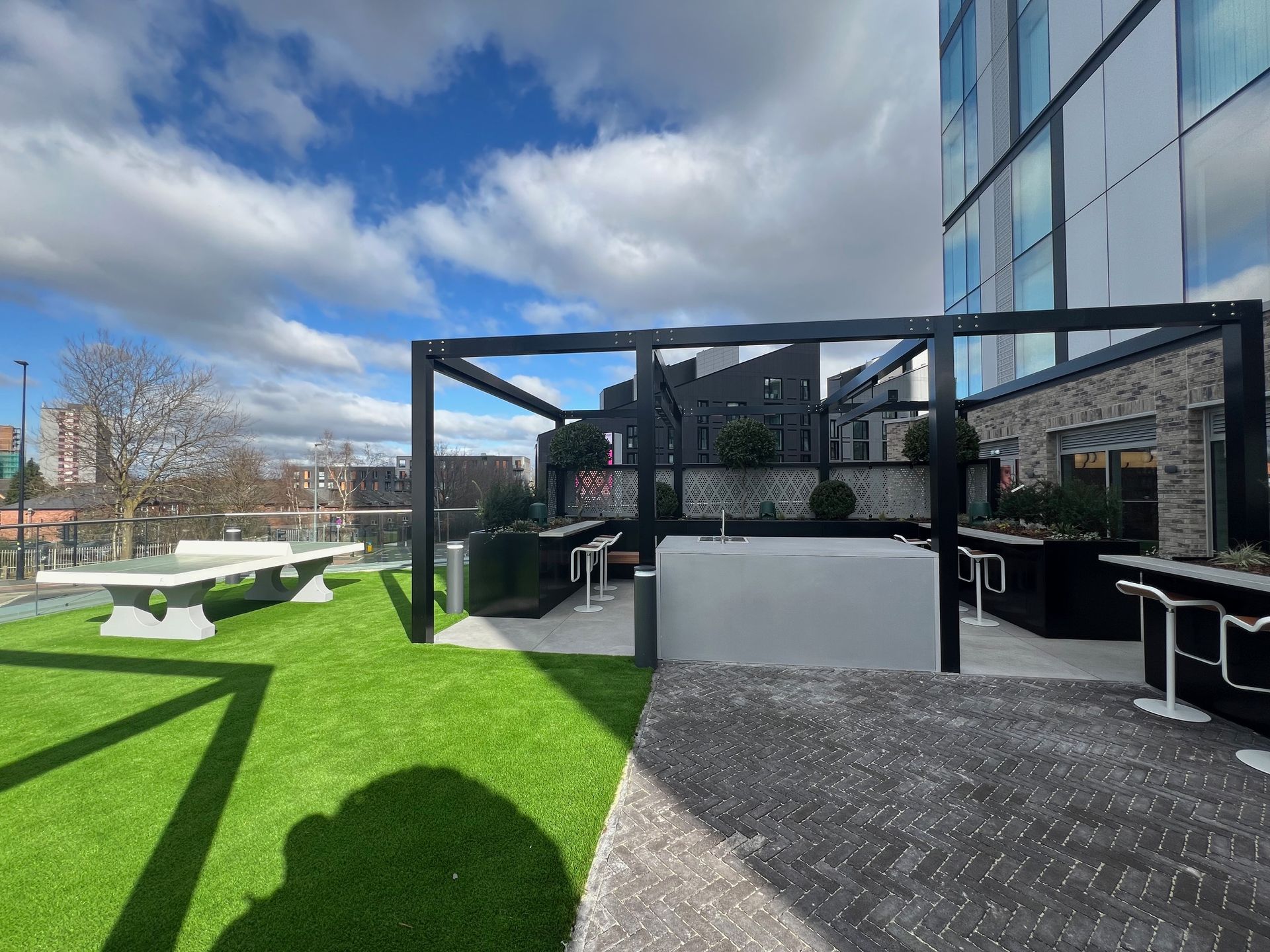 Rooftop patio with artificial turf, pergola, table tennis, and modern building under a cloudy sky.