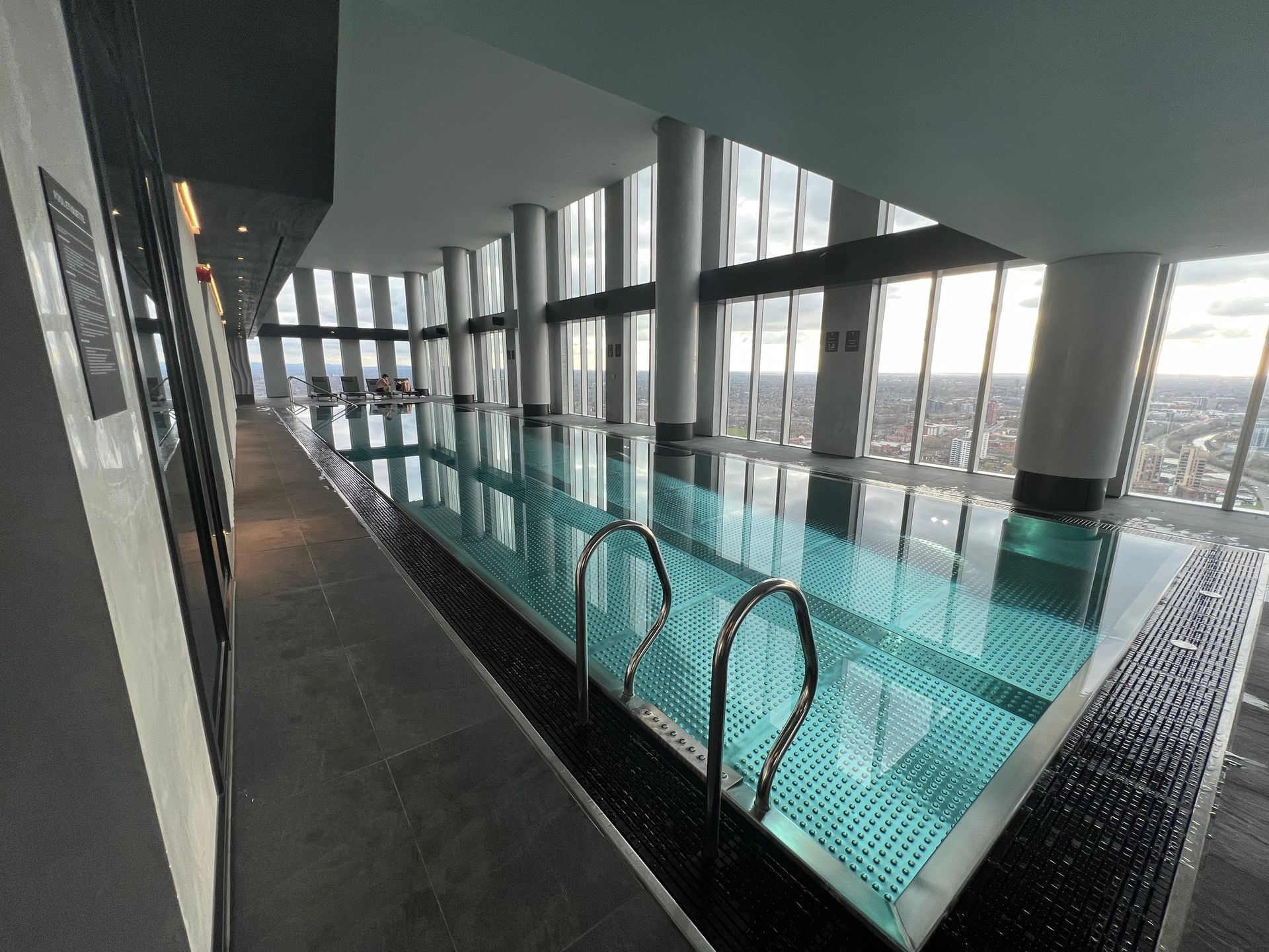 Indoor pool with city view, light blue water, black tiles, tall windows, and support columns.