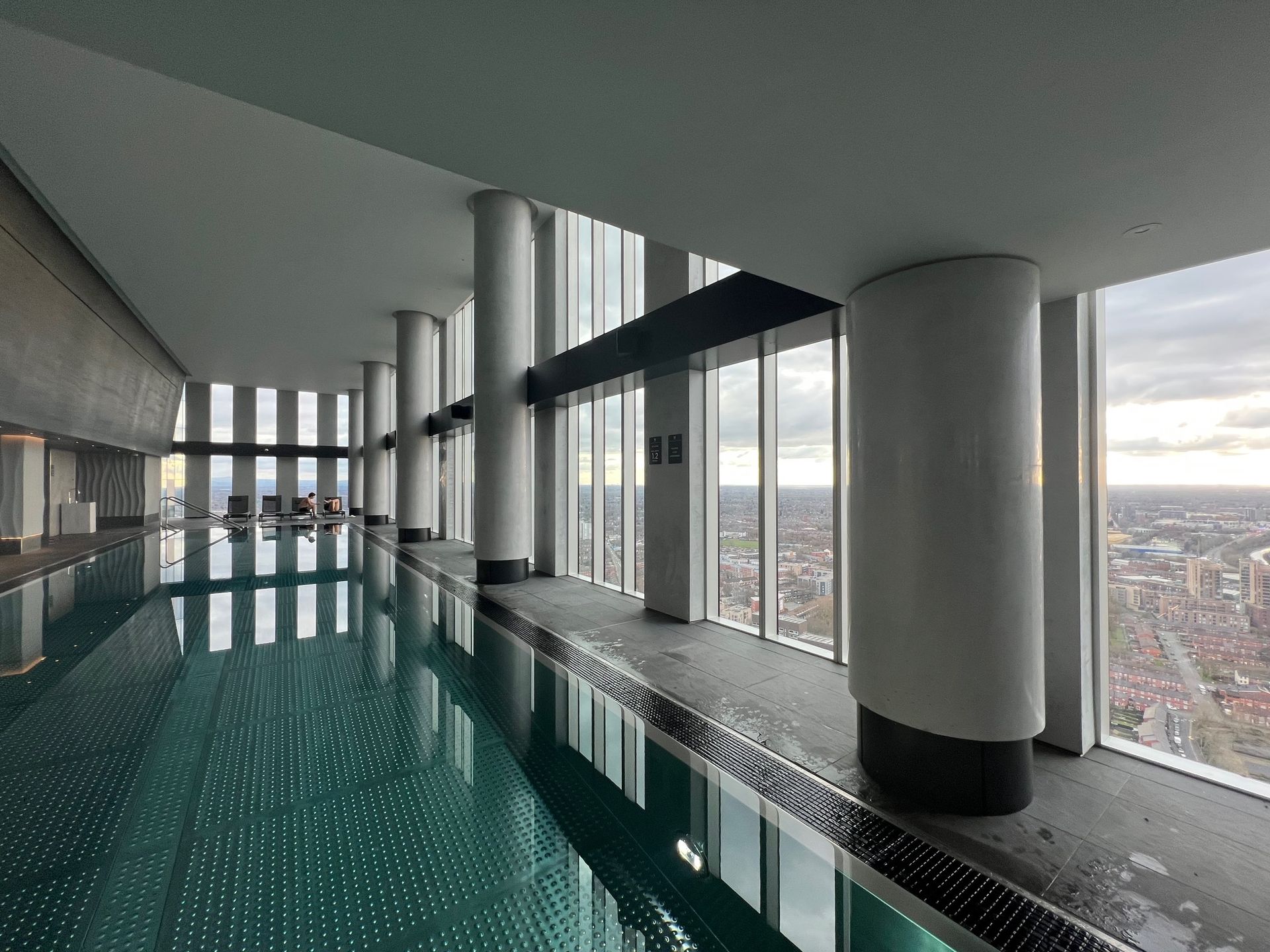 Indoor swimming pool with city view, white pillars, large windows, and reflective water.