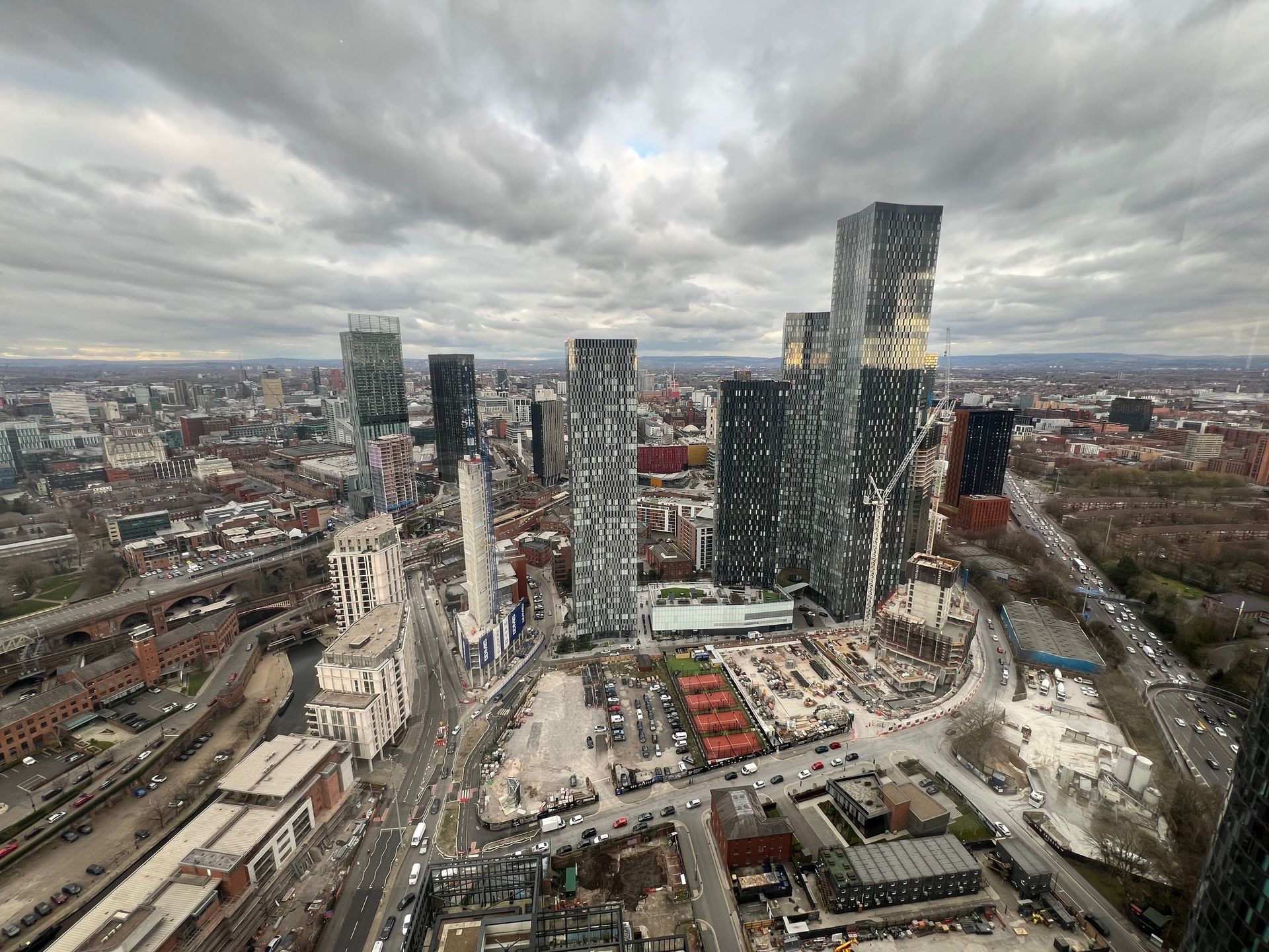 City skyline under cloudy sky, featuring tall modern buildings, streets, and industrial areas.