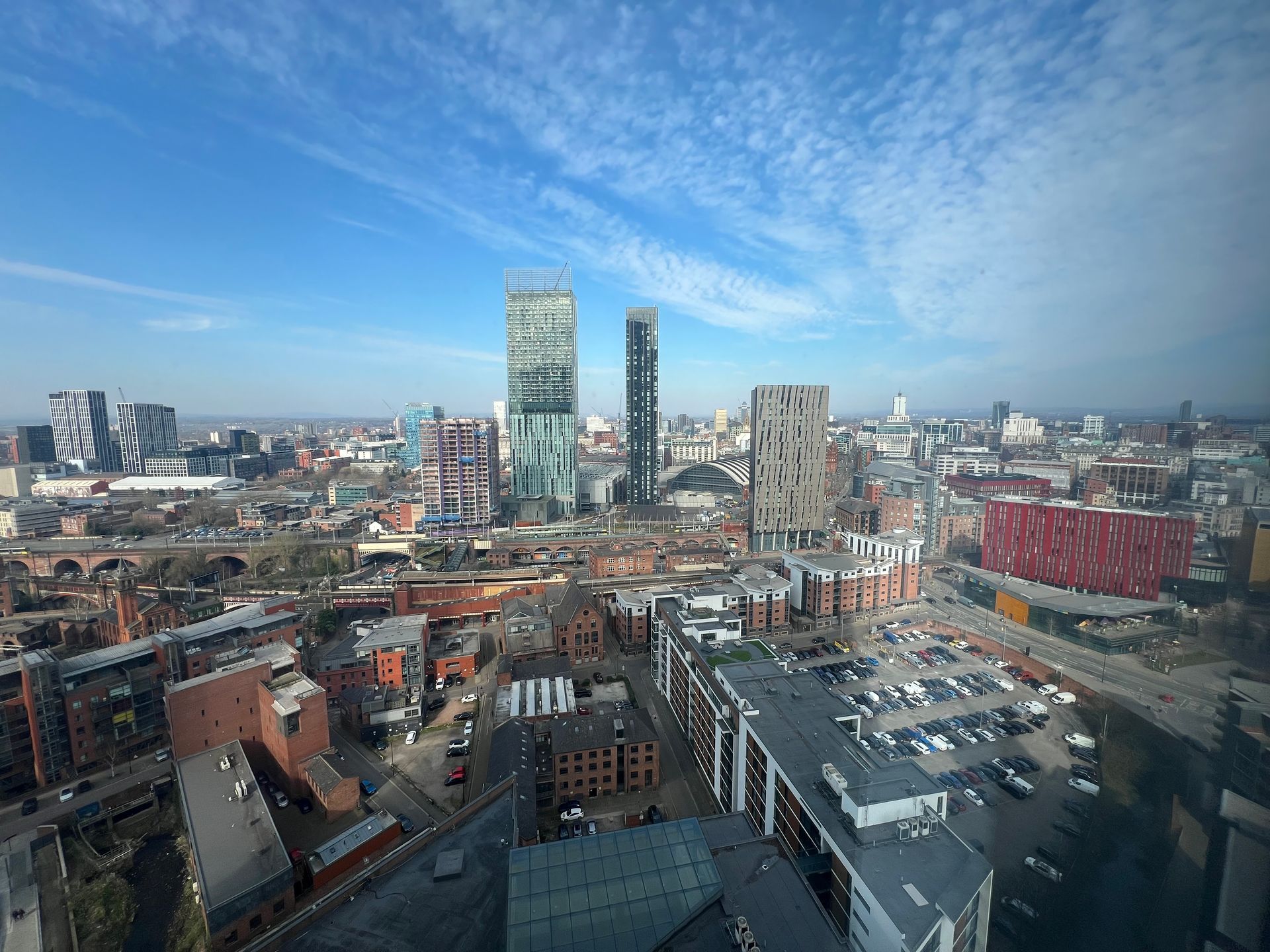 Cityscape of Manchester, UK, with modern skyscrapers under a blue sky with scattered clouds.