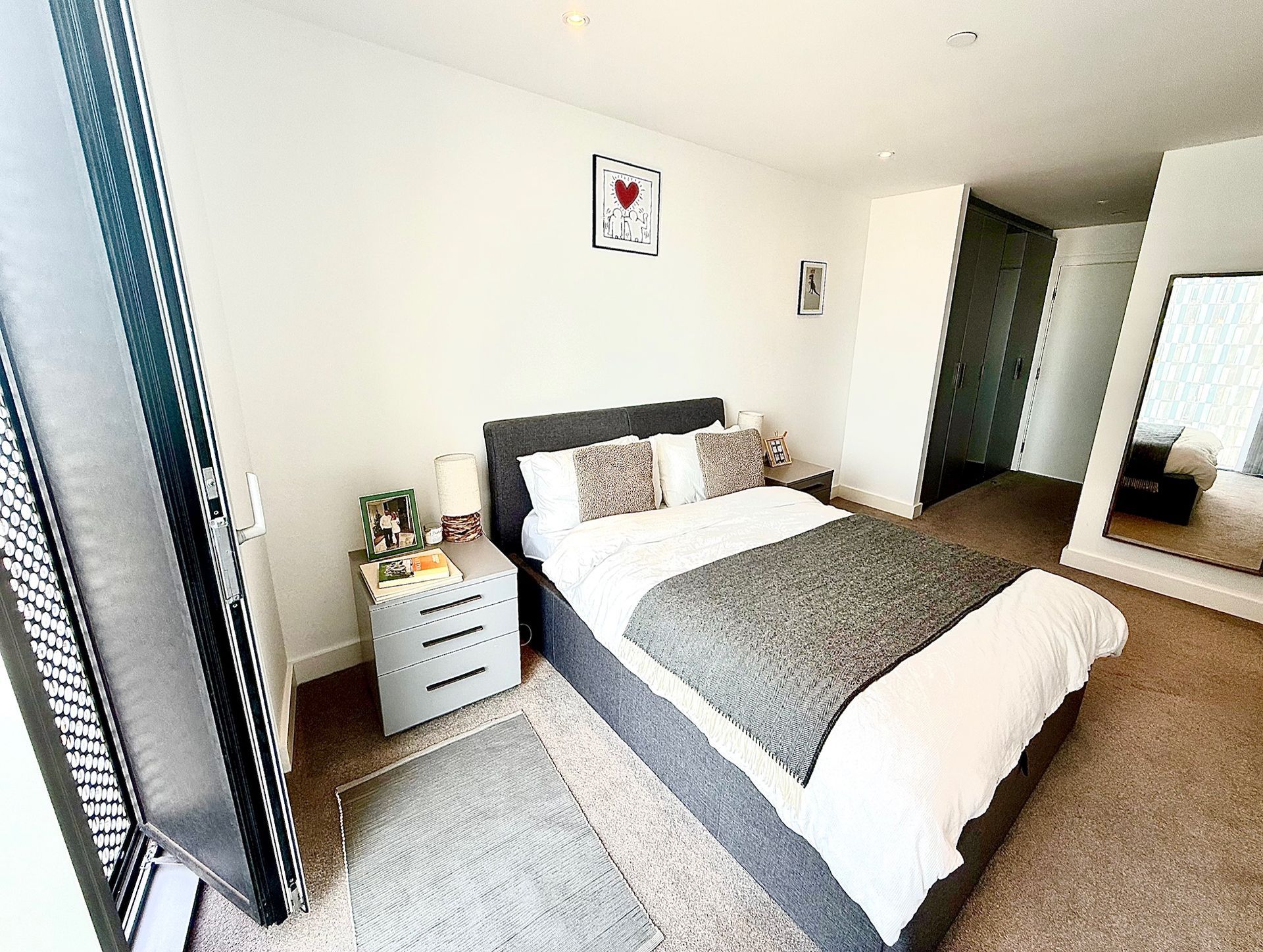 Bedroom with bed, nightstand, and large mirror. Window to the left.