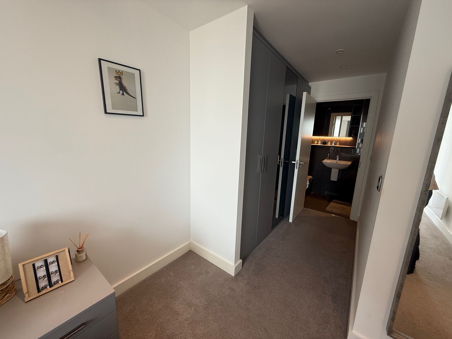 A narrow hallway with a light gray wall, leading to a bathroom. A framed picture hangs on the left wall.