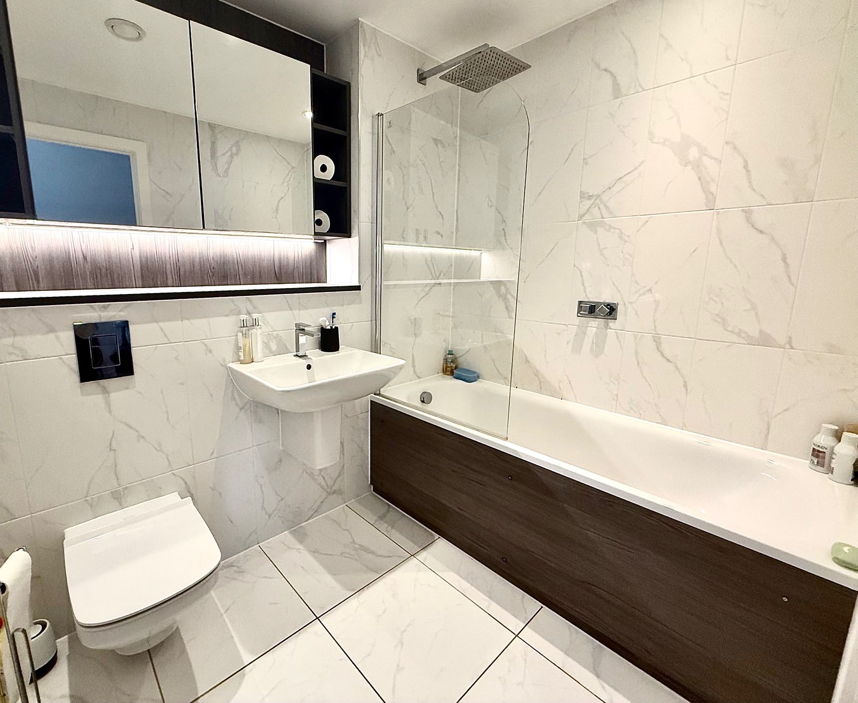 Modern bathroom with white marble-look tiles, a toilet, sink, bathtub, and glass shower screen.