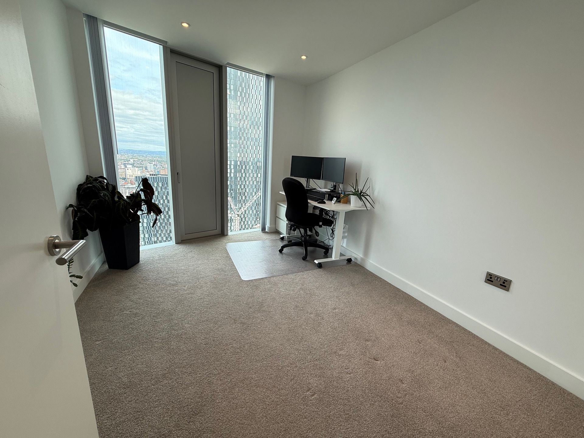 Empty room with a desk and monitors near a large window. A backpack sits near the door. Beige carpet.