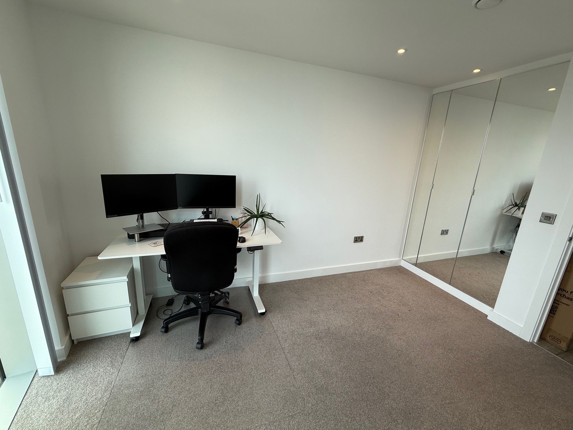 Home office with a white desk, two monitors, black chair, and mirrored closet.