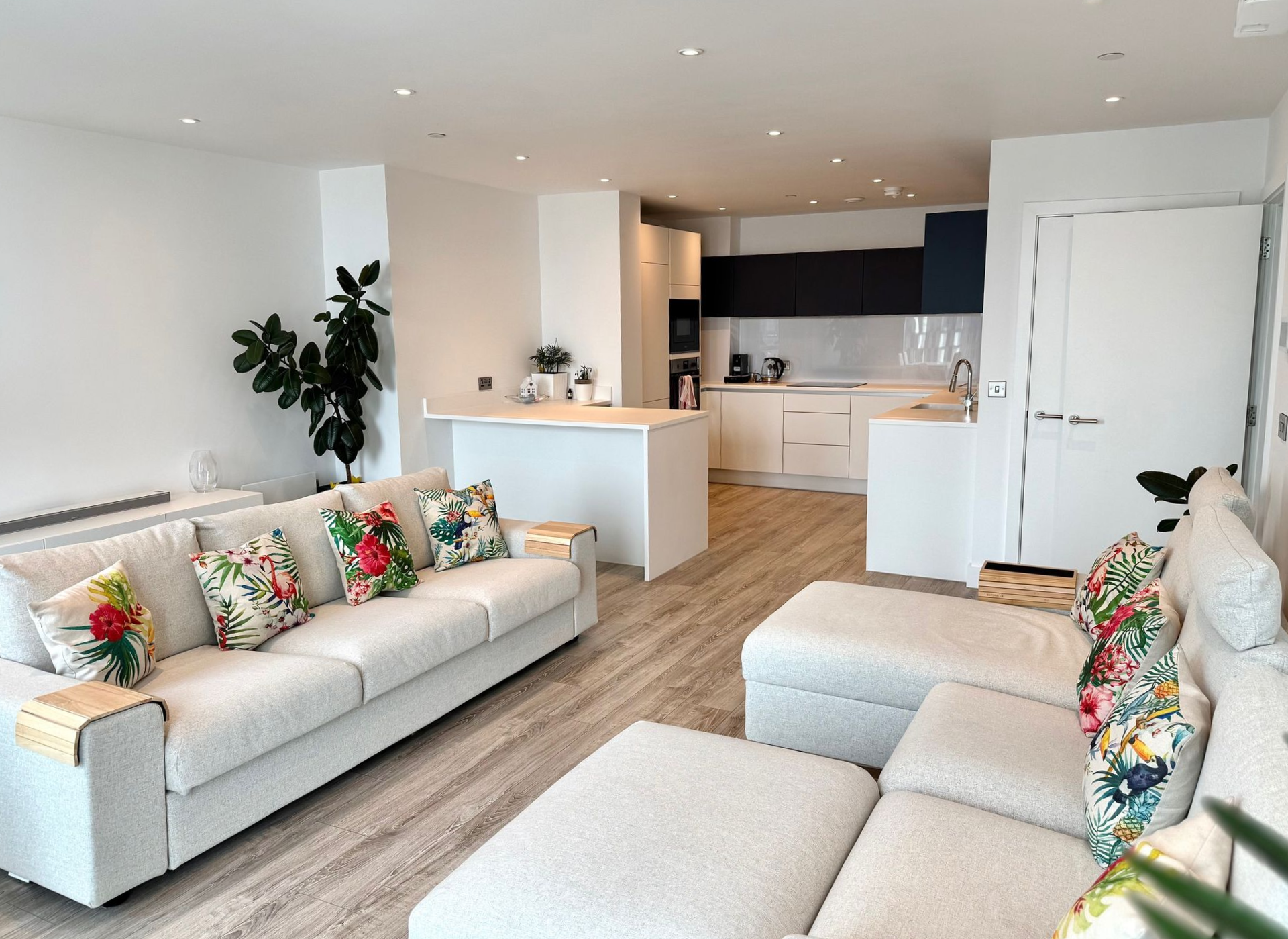 Living room with white sofas, floral pillows, light wood floors, and open kitchen.