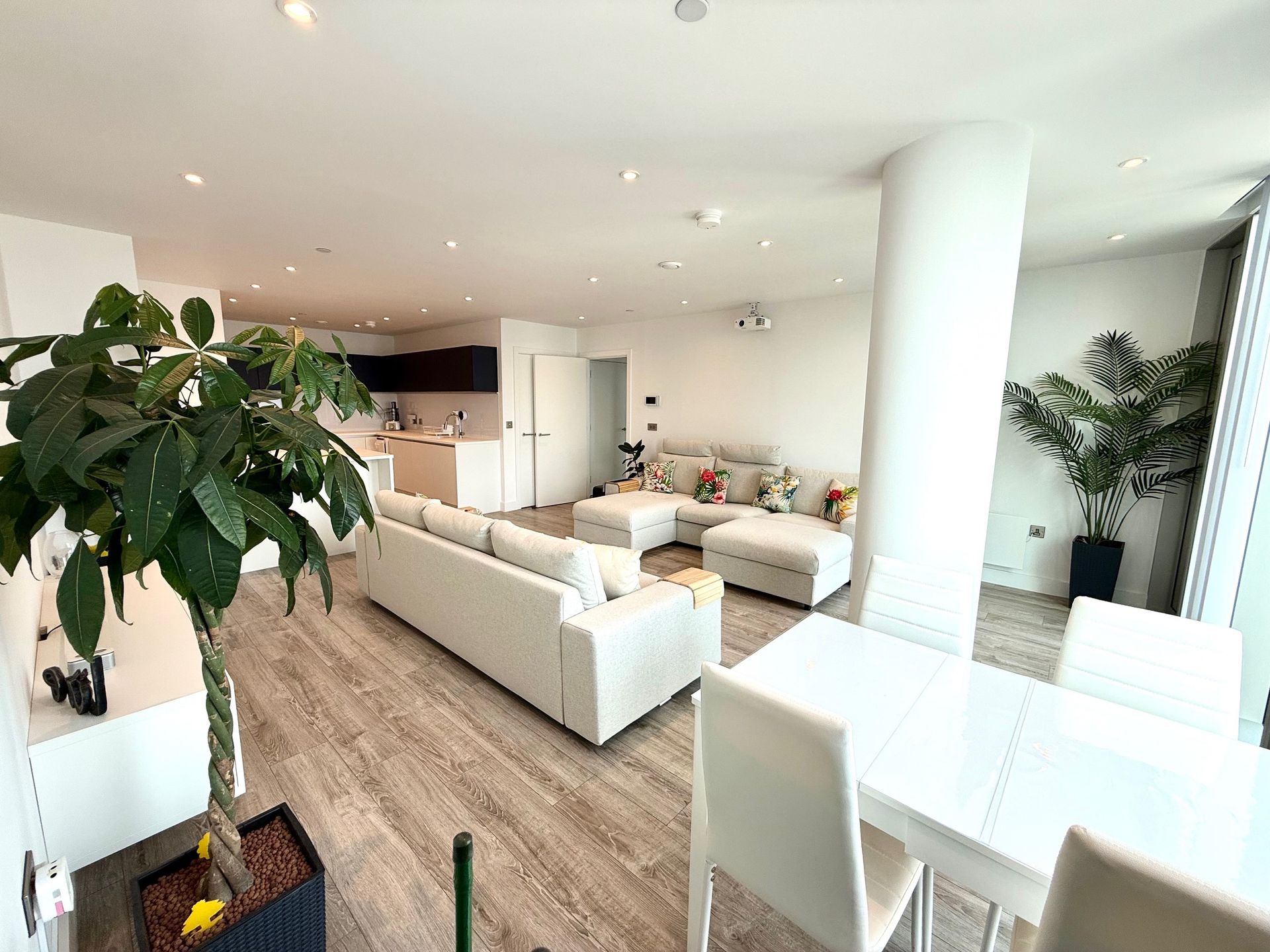 Bright living room with white sofas, dining table, and plants. Kitchen visible in the background.