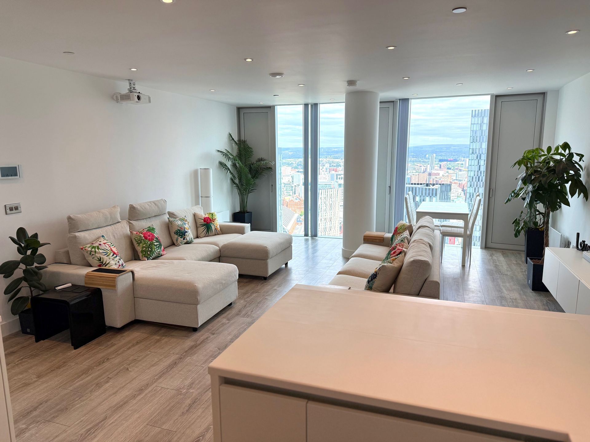 Modern living room with light-colored sectional sofas, large windows, city view, and plants.