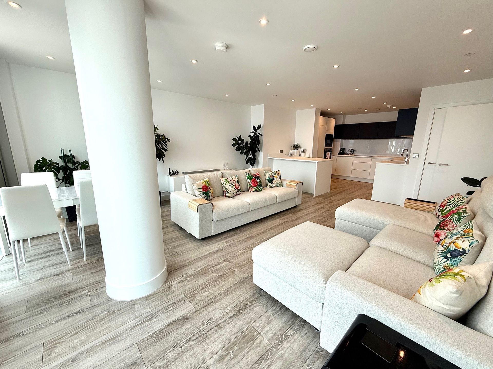 Spacious, modern living area with white furnishings and open kitchen. Includes dining, seating areas, and a large support pillar.