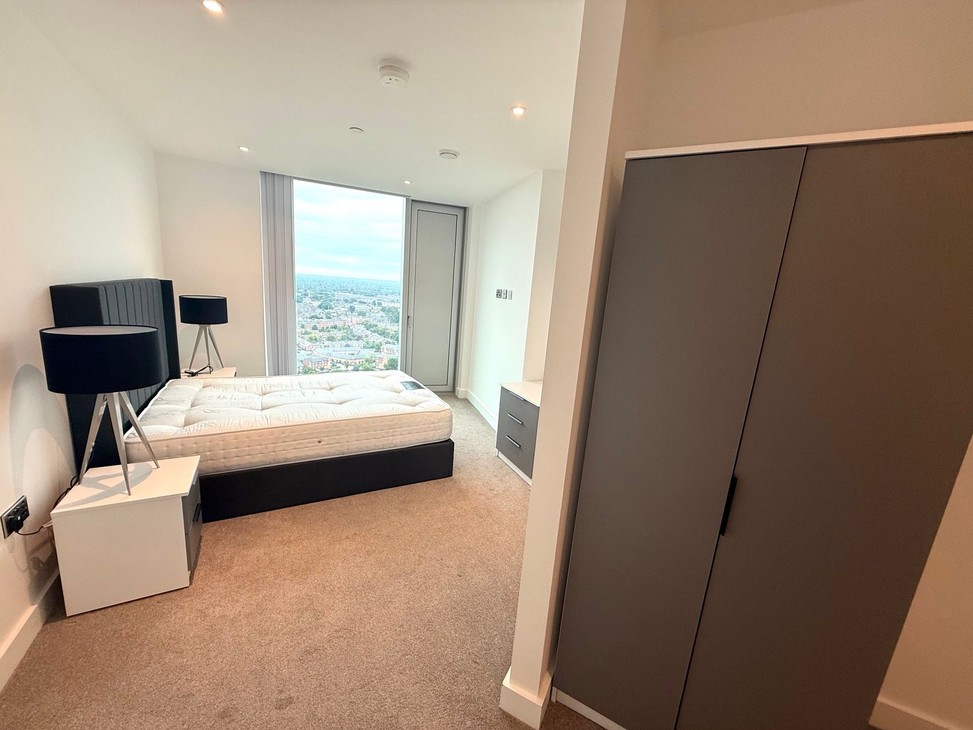 Bedroom interior: bed near window, bedside tables with lamps, gray wardrobe, and beige carpet.