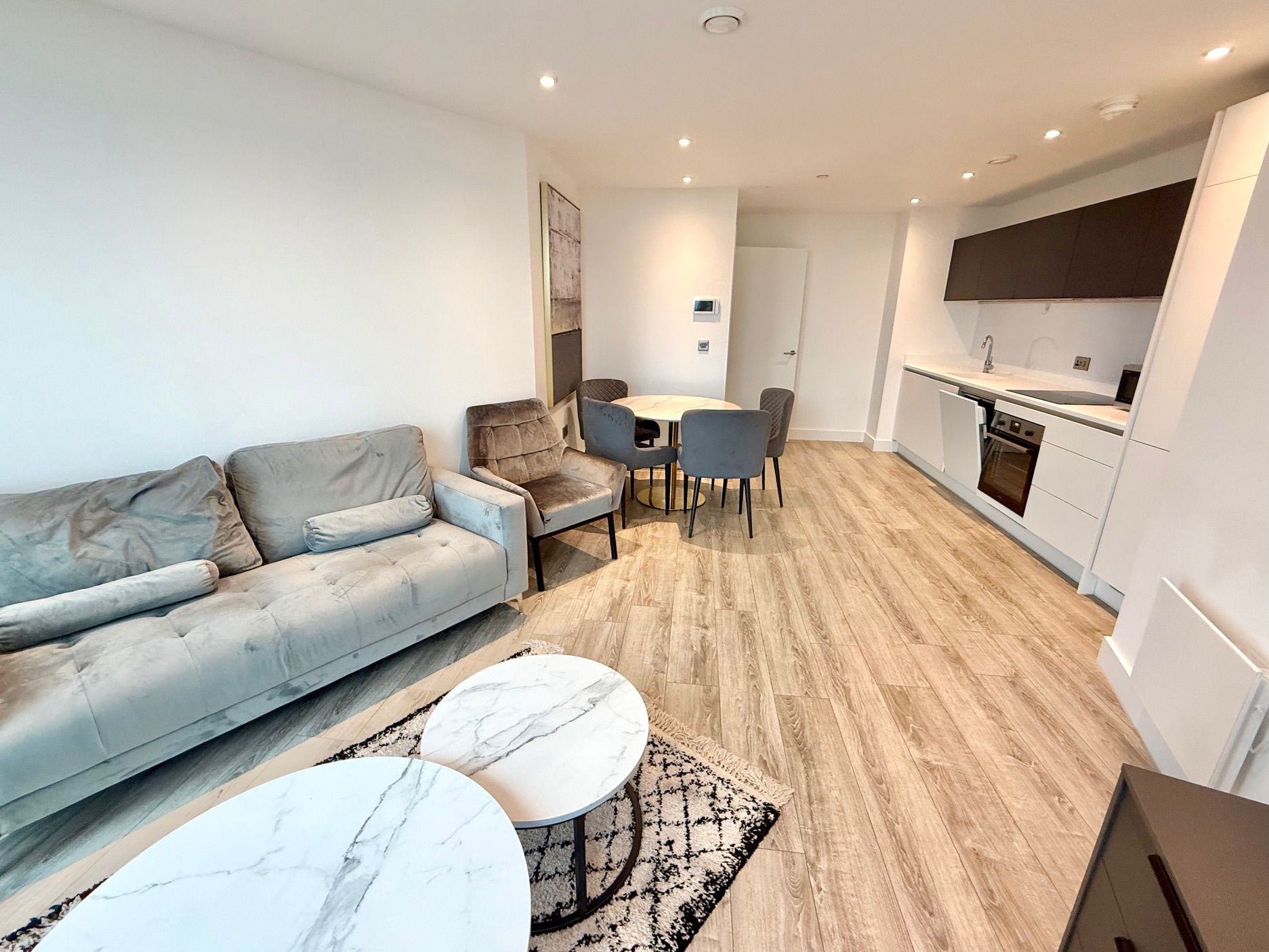 Modern living room with sofa, dining area, and open kitchen. Light wood floors, white walls.