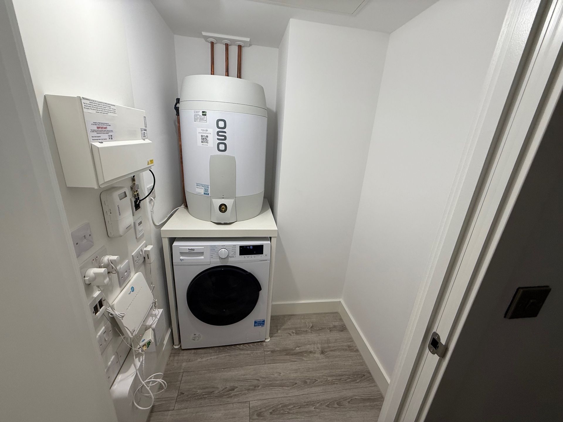 Small utility room with washing machine, water heater, and electrical panel. White walls, grey floor.