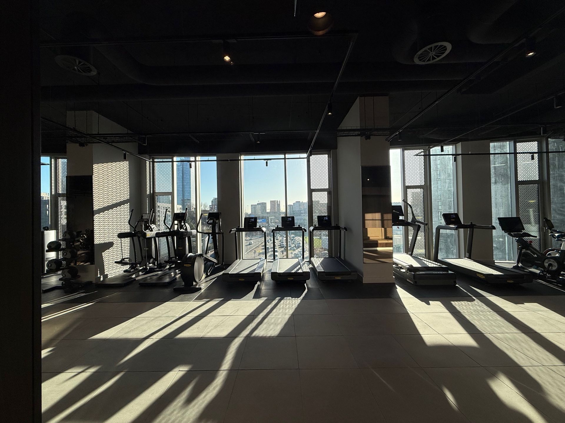 Brightly lit gym with treadmills and exercise equipment. Windows offer city views. Sunlight casts shadows.