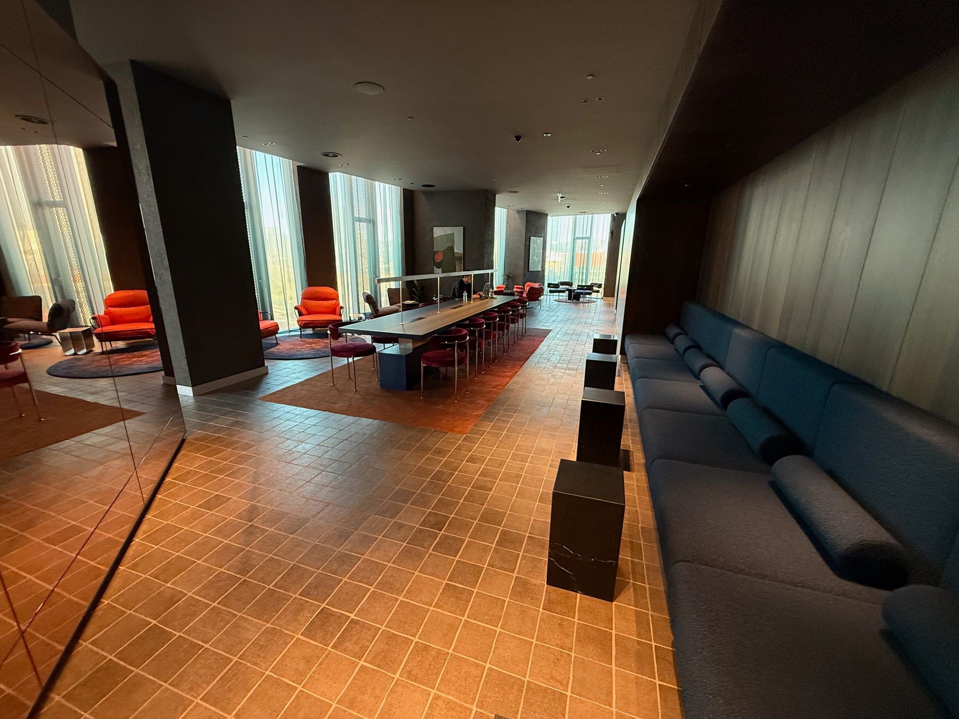 A modern lounge with a long table, sofas, and armchairs. Warm wood flooring and large windows.