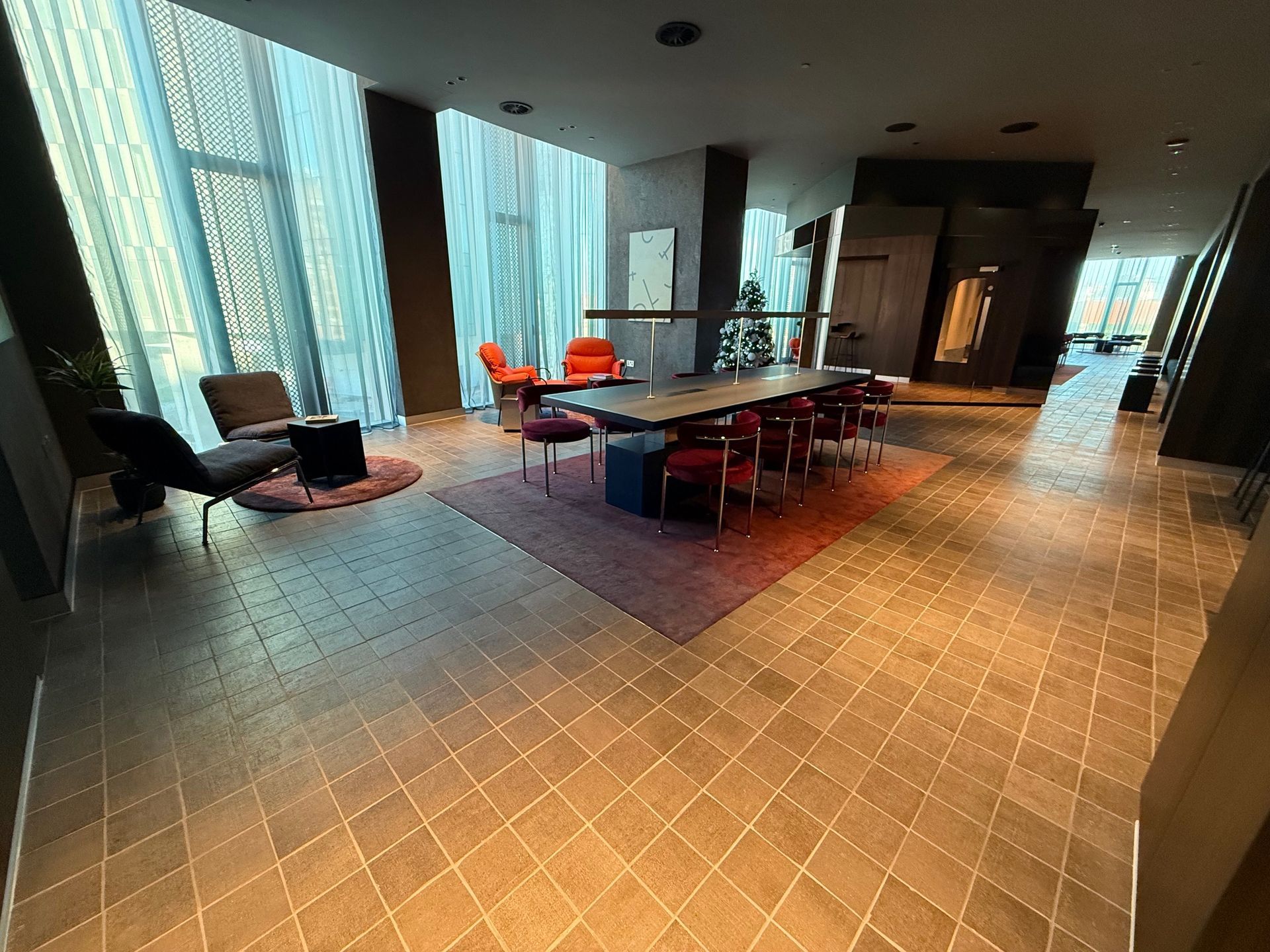 A modern office lobby with long table and chairs, armchairs on a rug, tall windows, and tiled floor.
