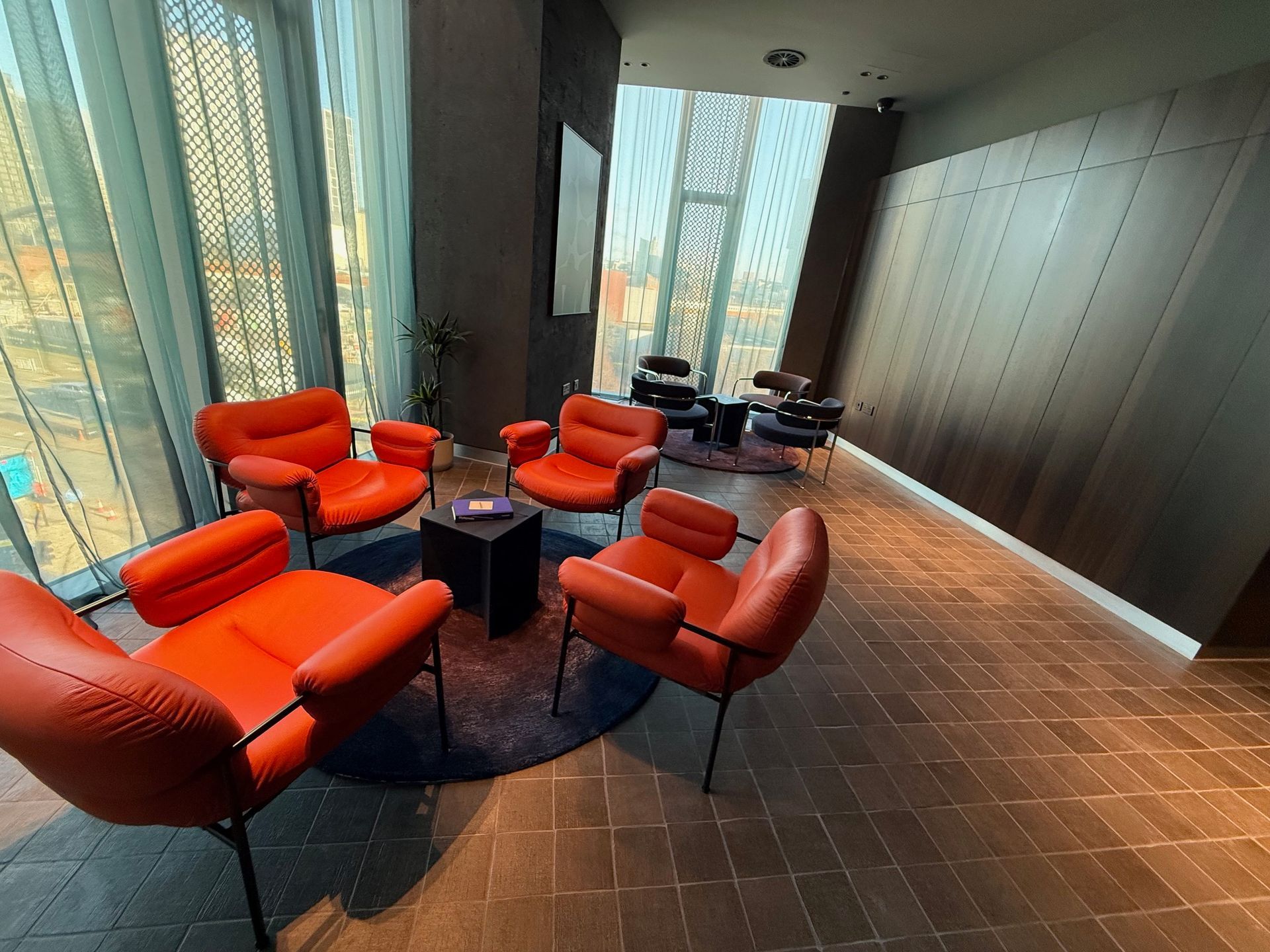 Orange chairs circle a black table on a rug, near windows and a wooden wall.