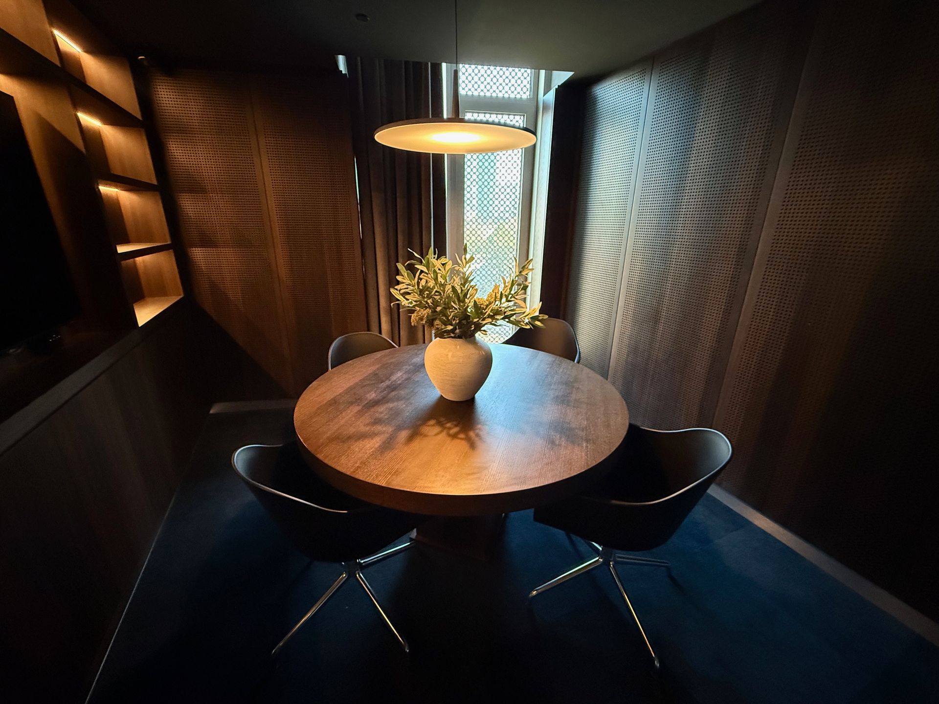 A dining area with a round table, chairs, flowers, and a pendant light. Dark wood walls and blue carpet.