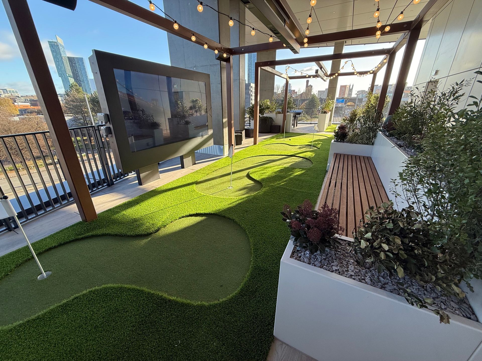 Rooftop putting green with artificial turf, plants, bench, and a large screen TV.