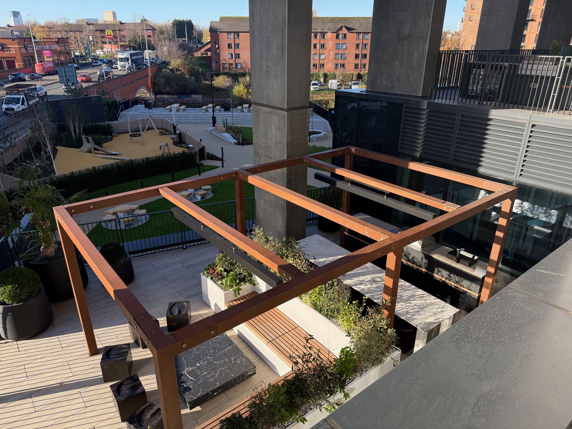 Outdoor terrace with brown pergola and greenery, overlooking a courtyard with buildings.