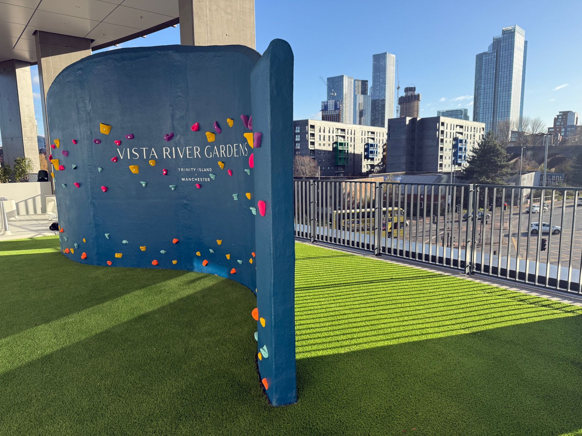 Blue climbing wall with colorful holds at Vista River Gardens; city skyline in background.
