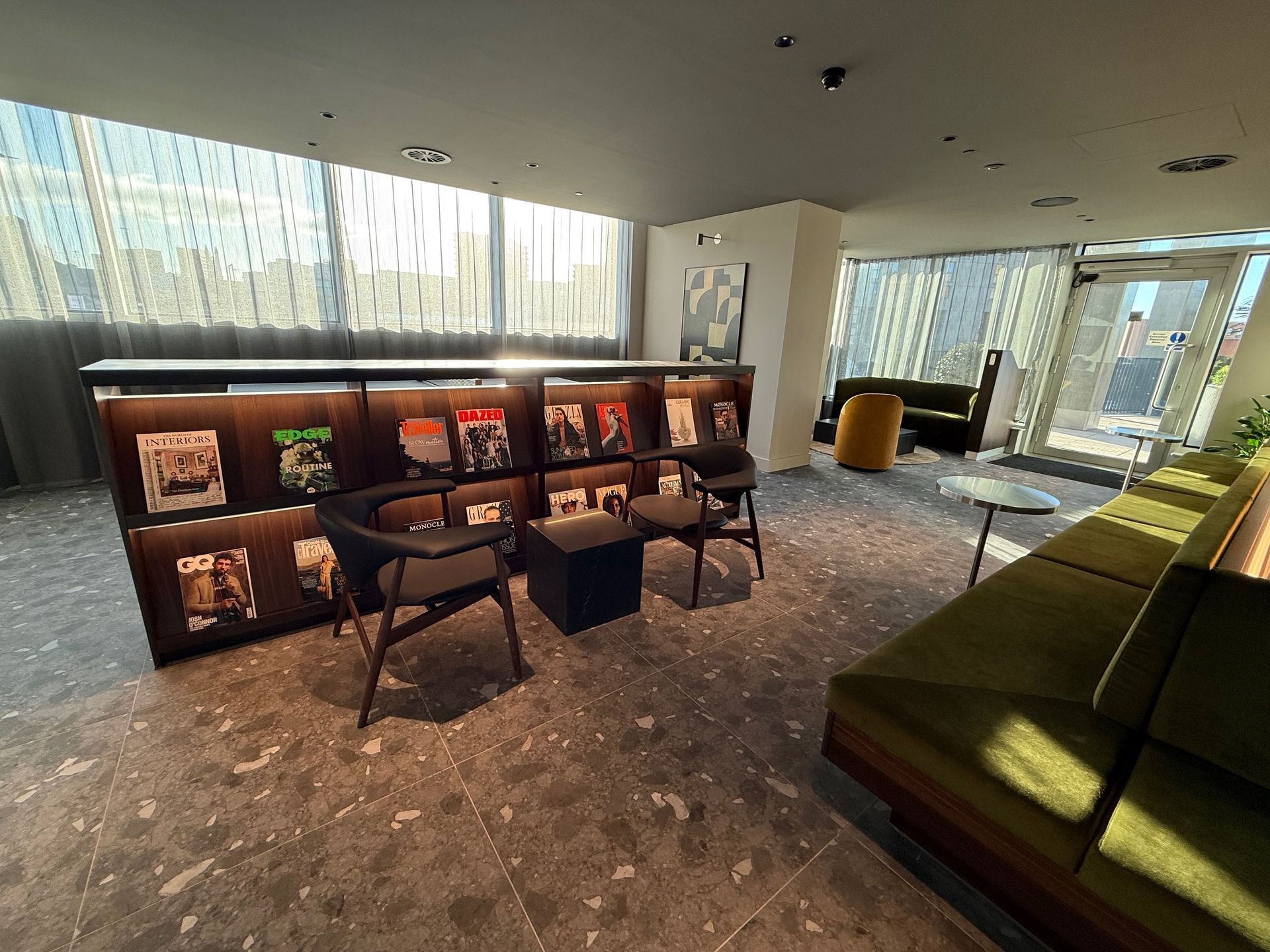 Lounge area with magazine display, seating, and large windows. Gray and brown color scheme, bright natural light.