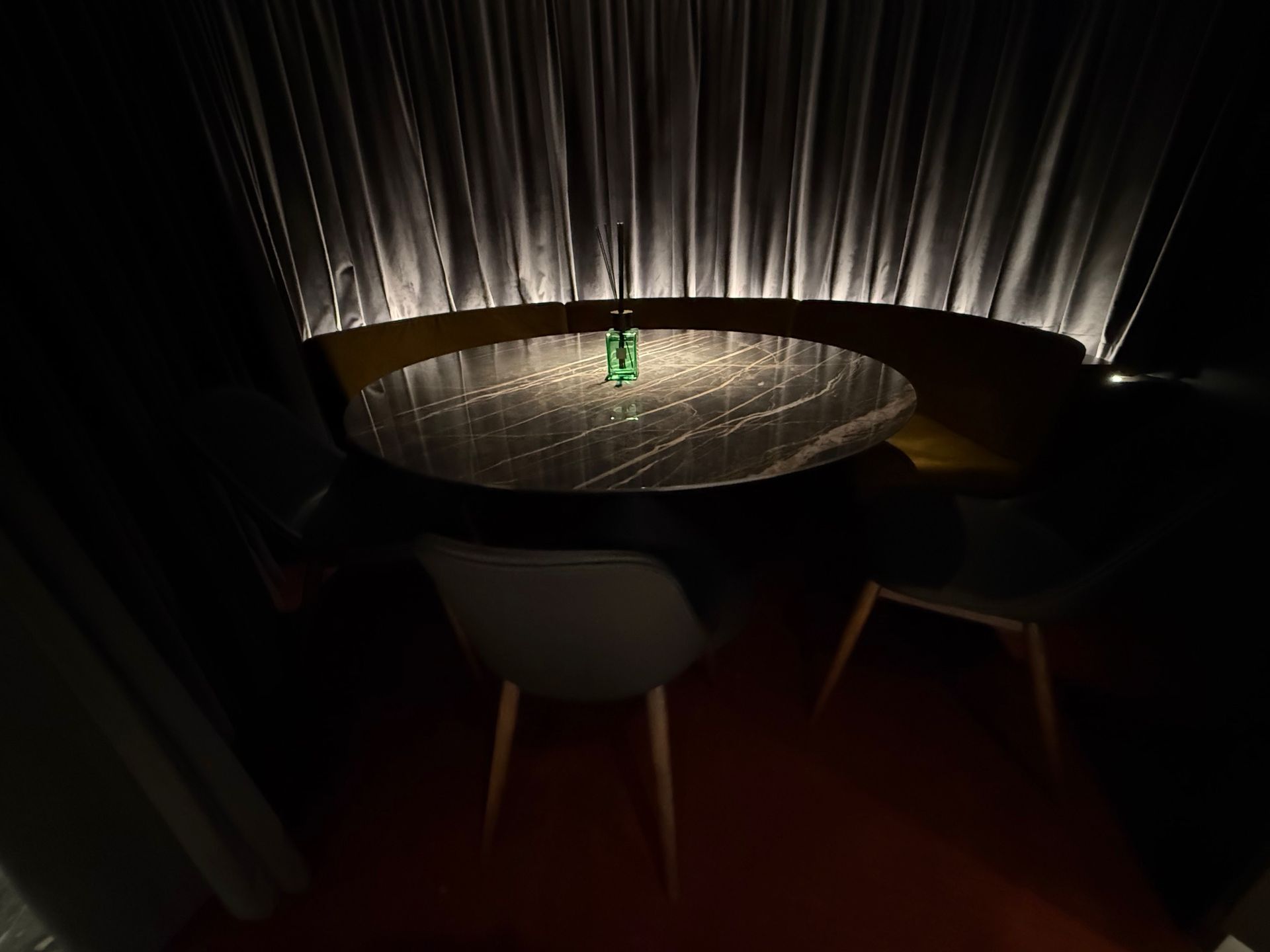 Dark interior with a round table, chairs, and a bottle. Backlit curtains provide a soft glow.