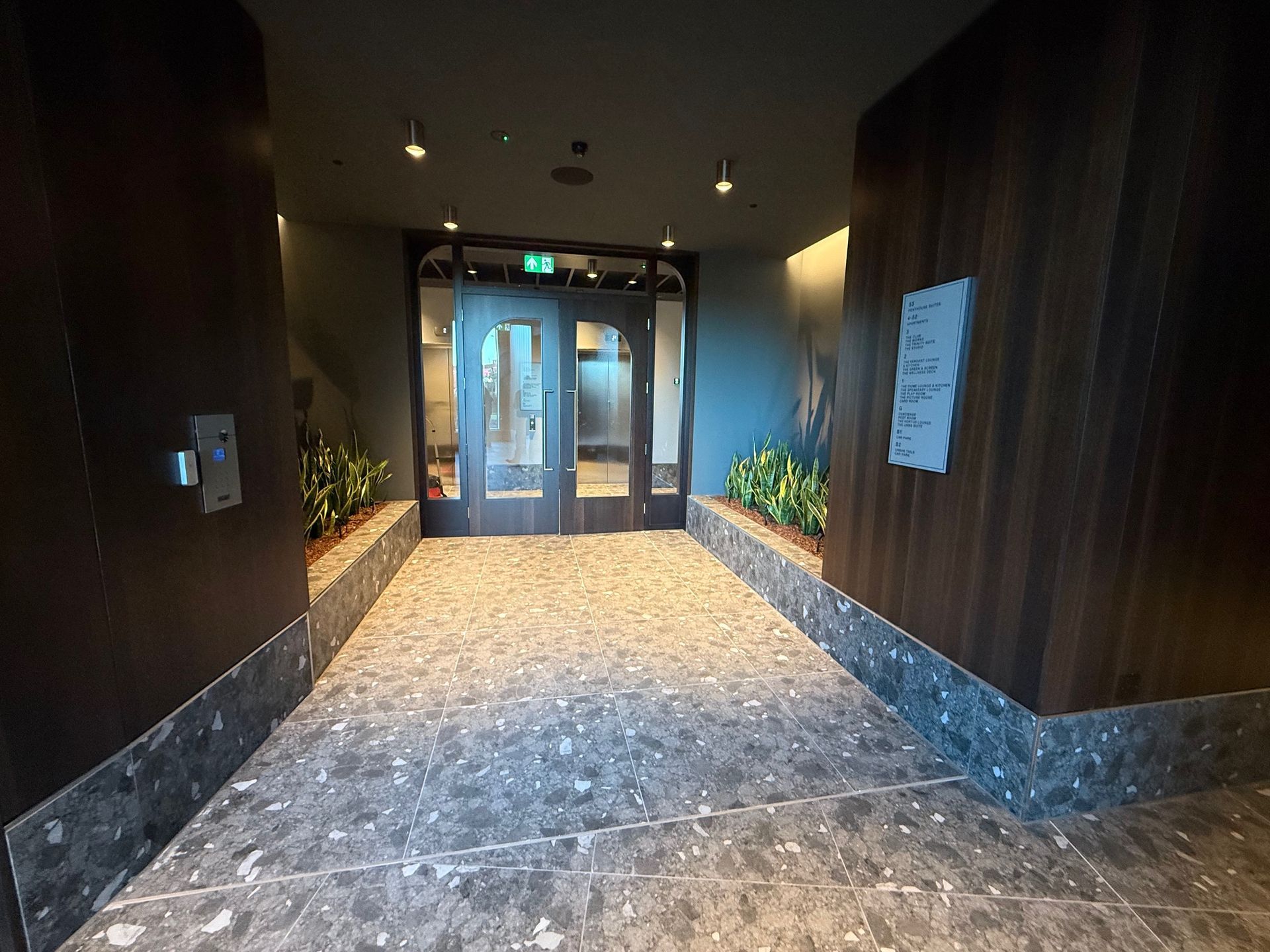 Hallway with glass doors at the end, leading outside. Dark wood and patterned flooring. Plants in stone planters.